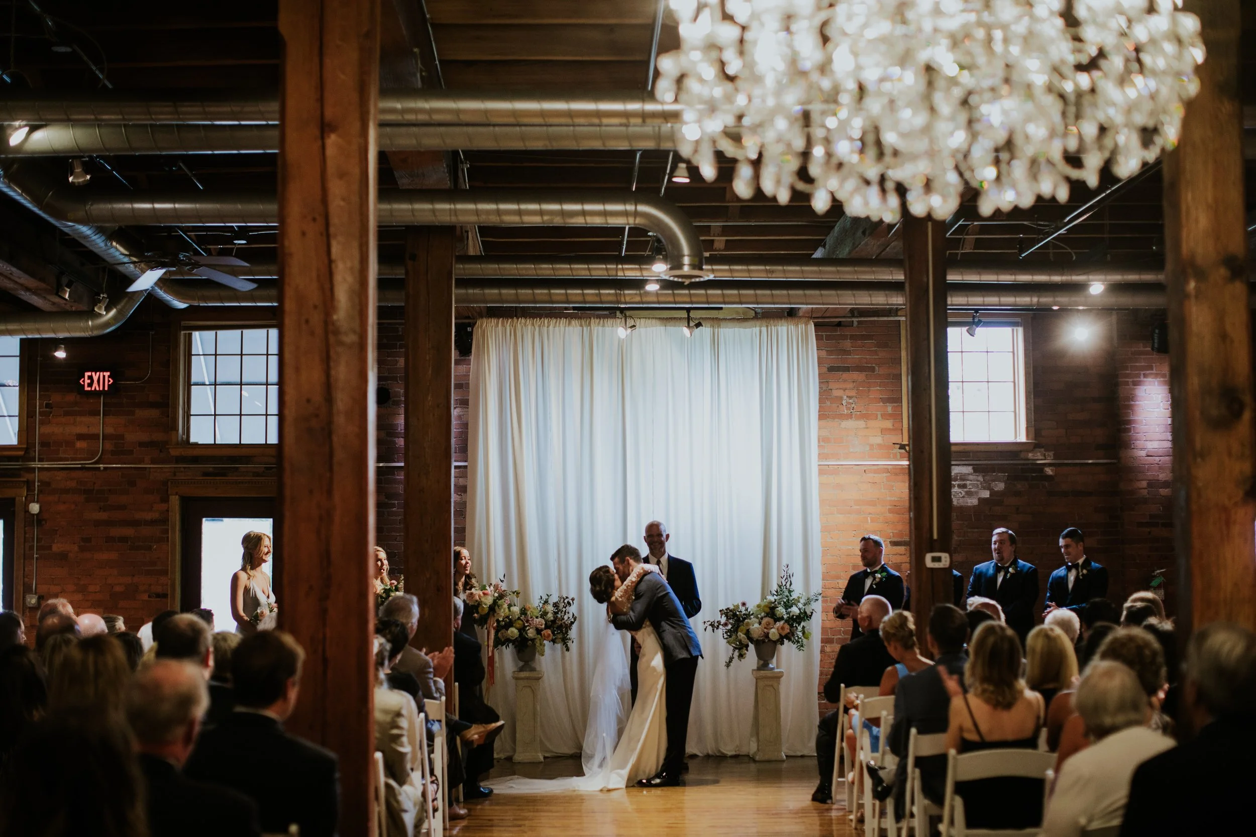 A wedding ceremony inside a rustic brick venue with exposed ductwork, large windows, and a chandelier. The bride and groom are kissing at the altar, surrounded by guests seated in white chairs.