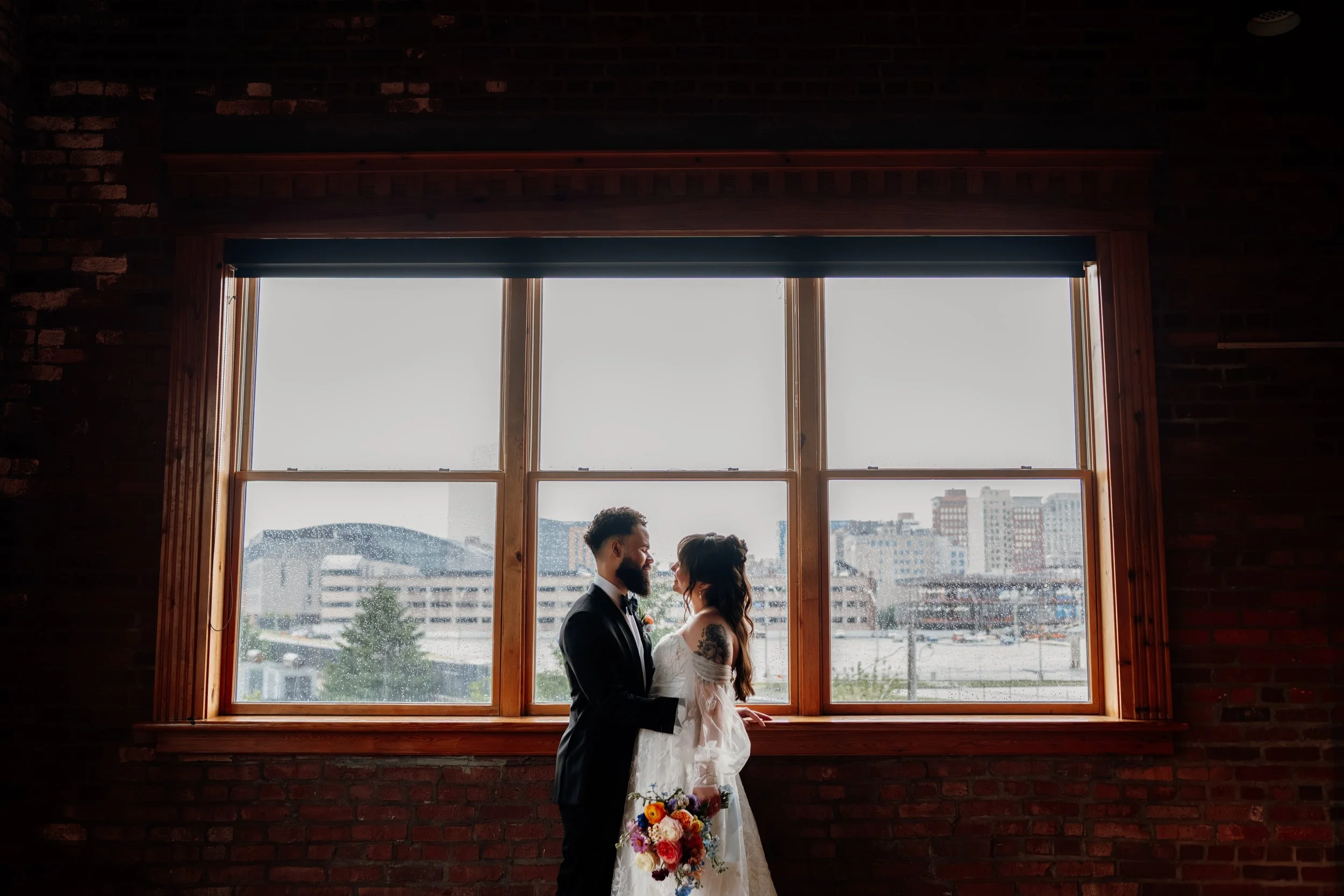 A couple in wedding attire standing close together by a large window, with an urban cityscape in the background on a cloudy day.