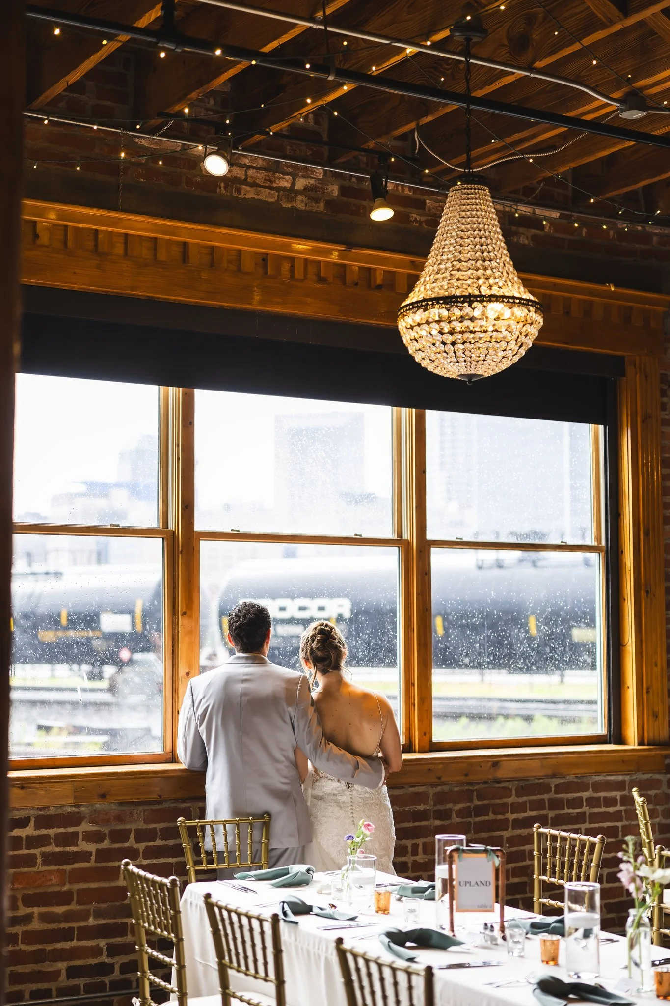 A couple stands by a large window with rain outside, offering a view of a train passing by. They are dressed for a wedding, with the woman in a white gown and the man in a grey suit. The interior features rustic brick walls, wooden beams, a large cha