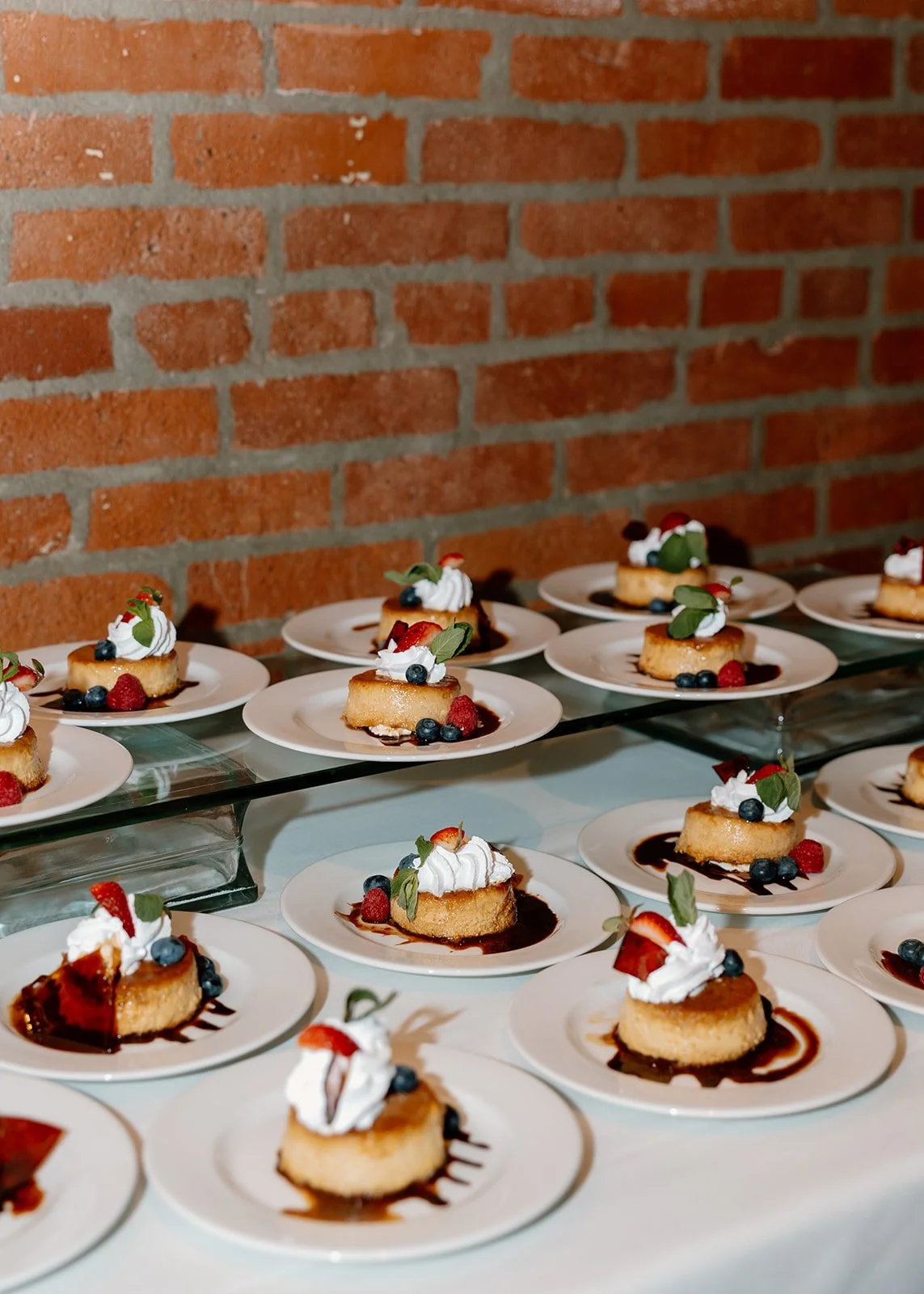 Several plates with mini cheesecakes topped with whipped cream, fruits, and mint leaves on a glass shelf against a brick wall.