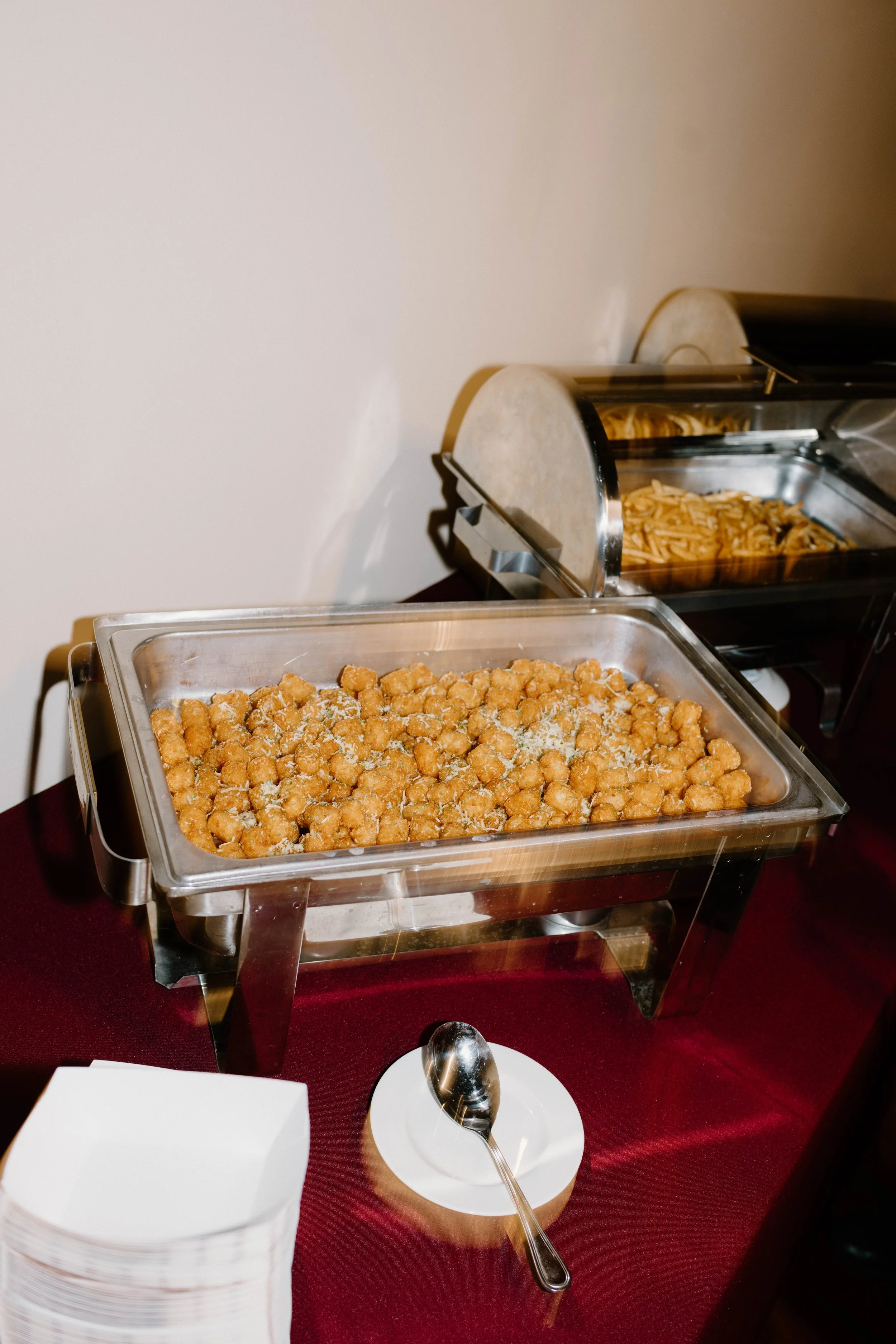 Chafing dish with fried chicken pieces, trays with pasta, and empty white bowls on a purple tablecloth, buffet-style setup.