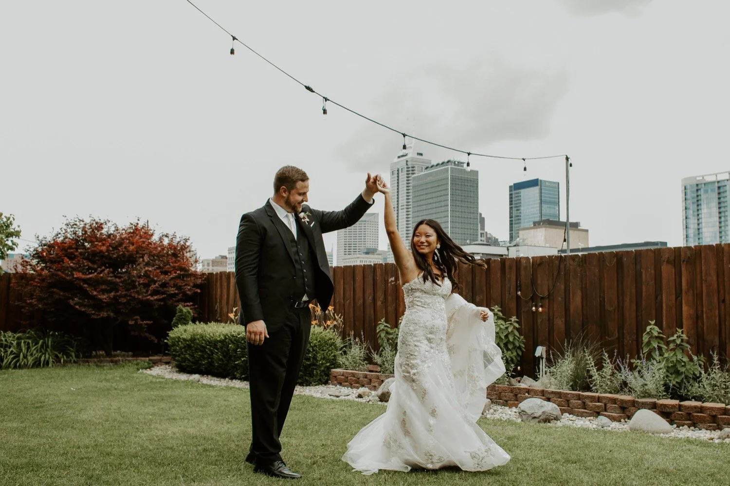 A couple dancing in a backyard with city buildings in the background. The woman is in a white wedding dress, and the man is in a black suit. The woman is smiling and holding the man's hand as he twirls her.