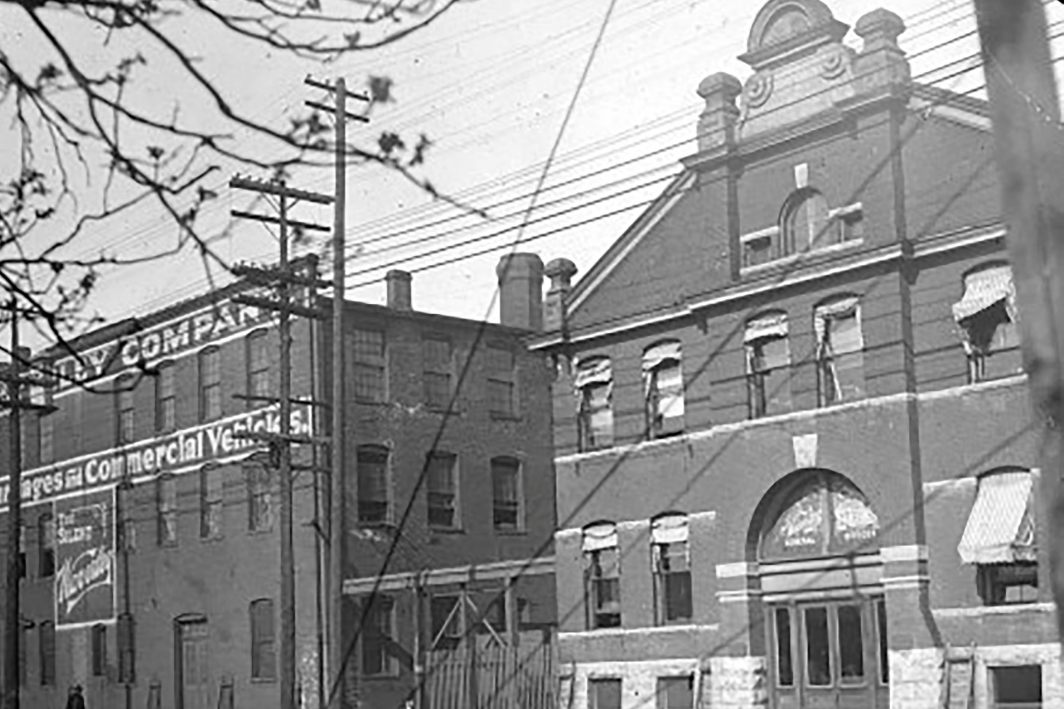 Black and white photograph of a city street scene with multi-story buildings, utility poles, and overhead wires. One building has a visible sign with the text 'Present Commercial Ve...' and decorative architectural features on the roof. Bare tree branches are in the foreground.
