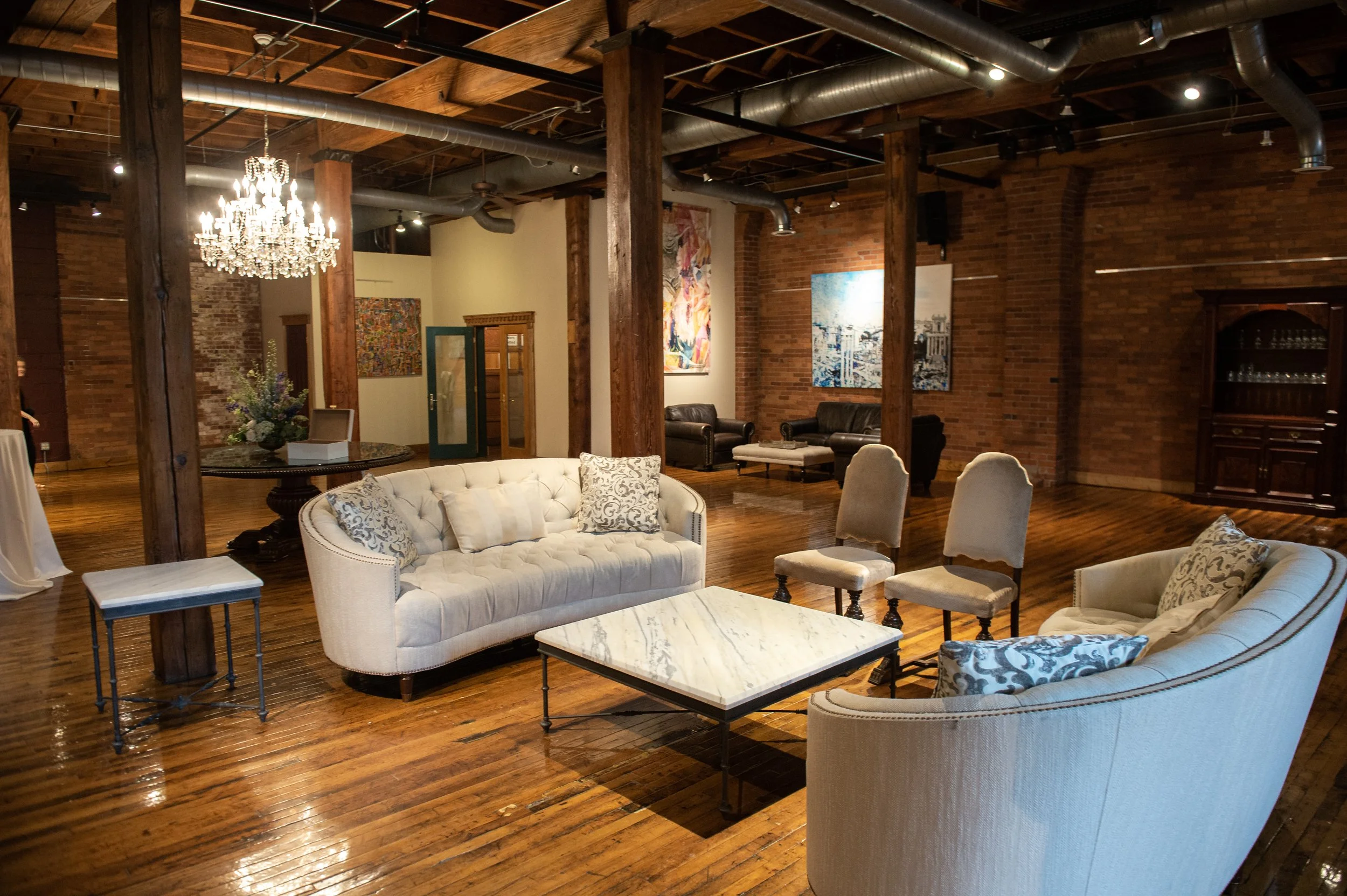Interior of an industrial-style living room with brick walls, wooden beams, a chandelier, multiple art pieces, and comfortable seating including a white tufted sofa and armchairs.
