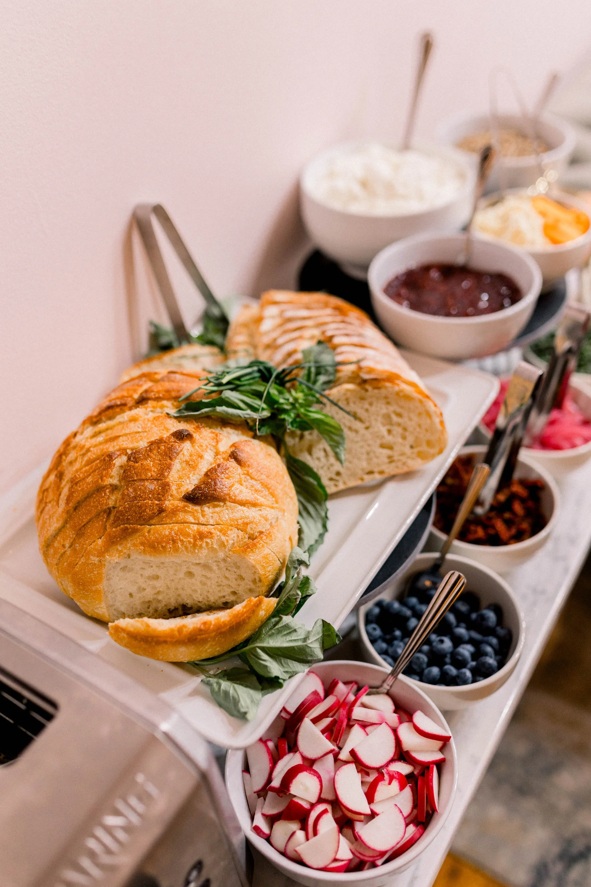 A buffet table with sliced bread, fresh fruit, and assorted breakfast toppings including yogurt, jam, blueberries, radishes, and other condiments.