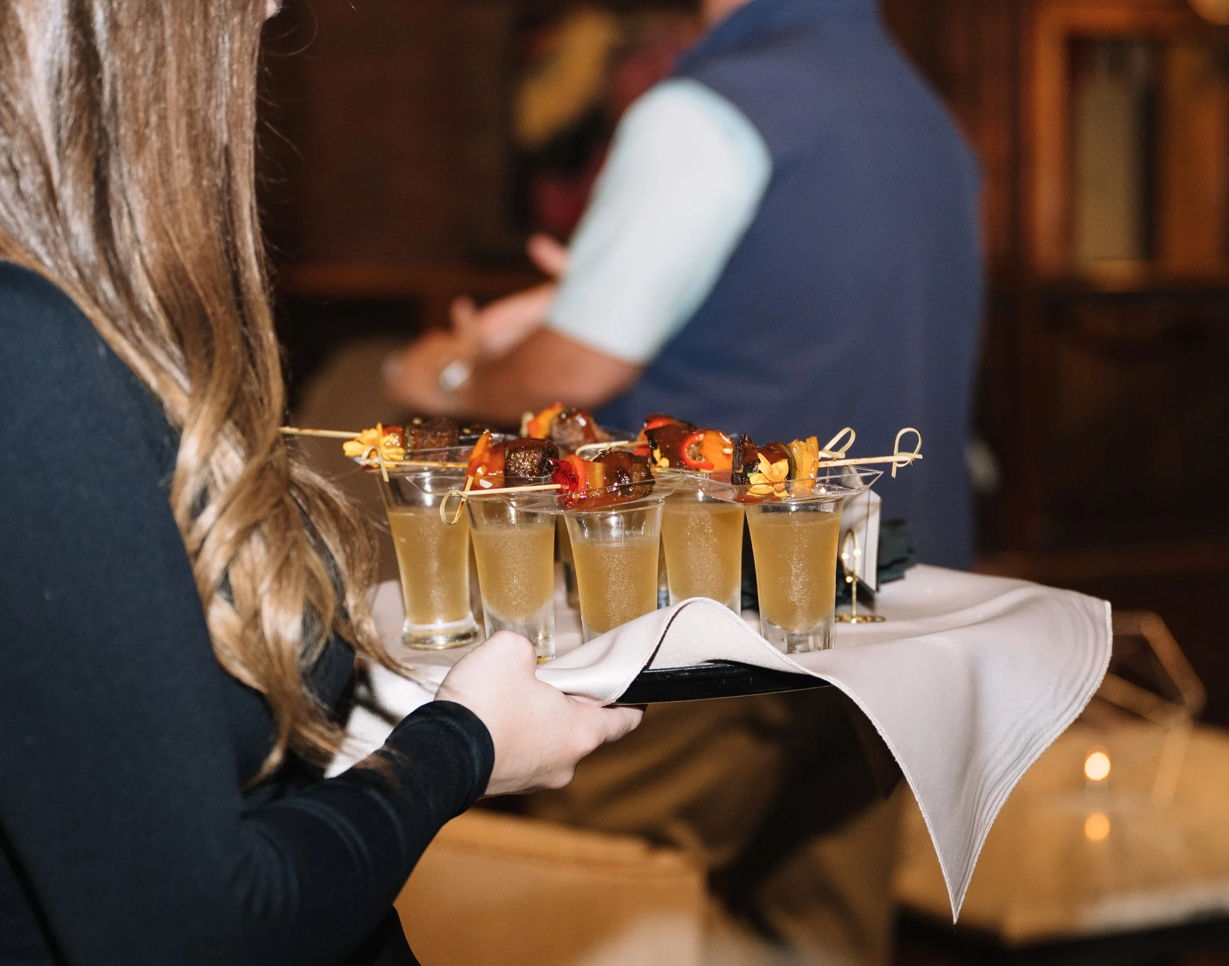 A person with long brown hair carrying a tray of small glasses filled with a light-colored drink and topped with skewers of grilled meat and vegetables at a social event, with warm lighting in the background.