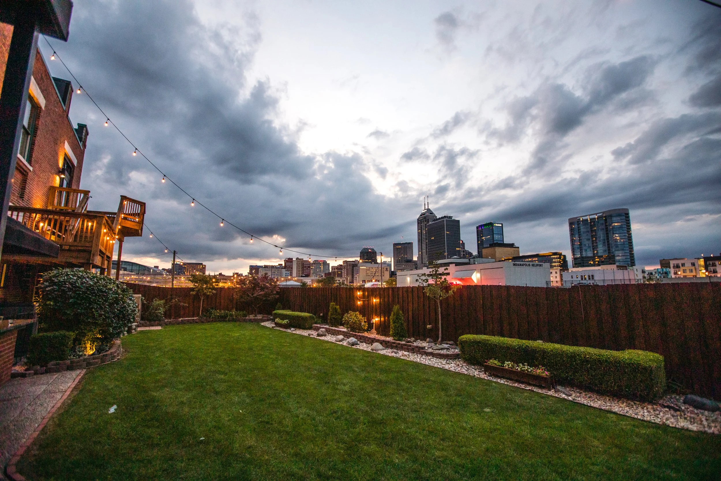 A backyard with a neatly manicured lawn, plants, and bushes, enclosed by a wooden fence. Elevated wooden decks with string lights are on the left, and a city skyline with tall buildings and a cloudy sky is in the background.