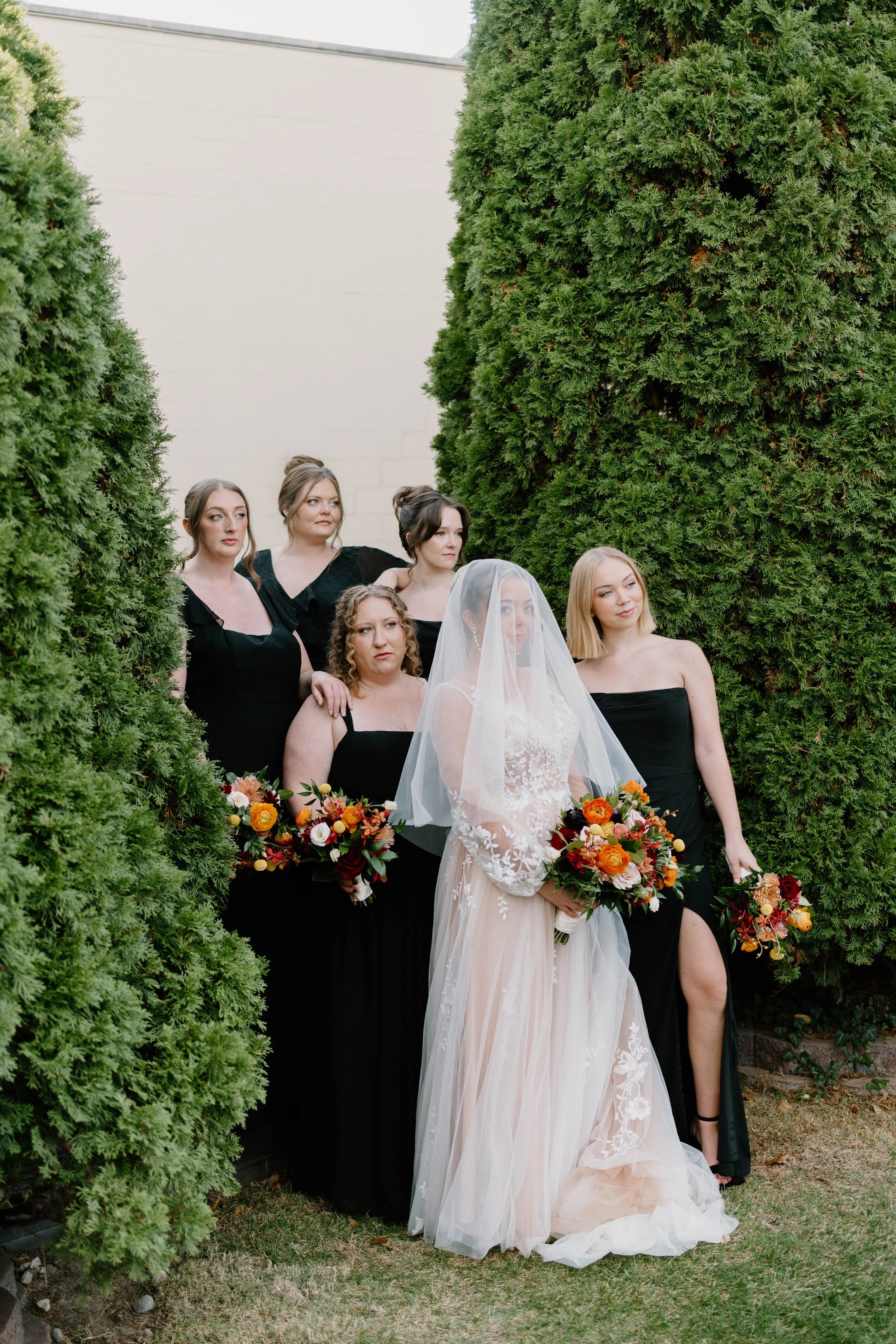 A bride and five bridesmaids standing outdoors in front of green trees, with the bride in a white gown and veil holding a bouquet of flowers, and the bridesmaids in black dresses holding similar bouquets.