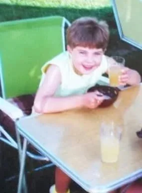 A young boy sits at a table outdoors, holding a remote control in one hand and a glass of orange juice in the other, smiling at the camera.