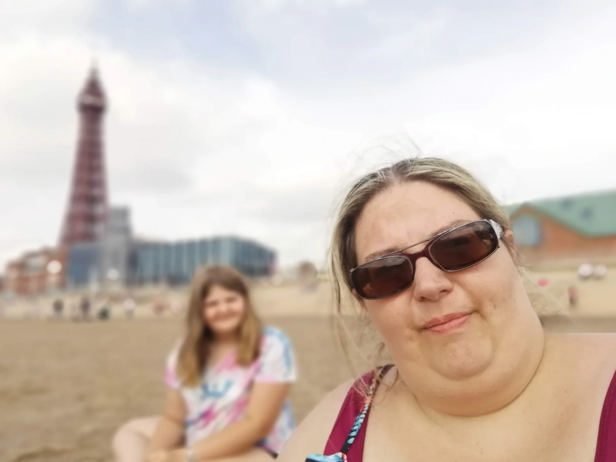 A woman with sunglasses smiling in the foreground, with a young girl in a colorful swimsuit standing in the background, at a beach with a blurred Eiffel Tower in the distance.