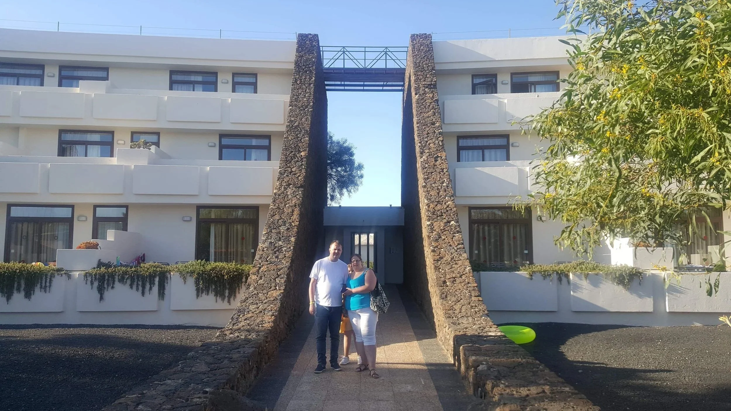 A man and a woman standing together in front of a modern white building with multiple balconies, flanked by stone walls and lush greenery, under a clear blue sky.