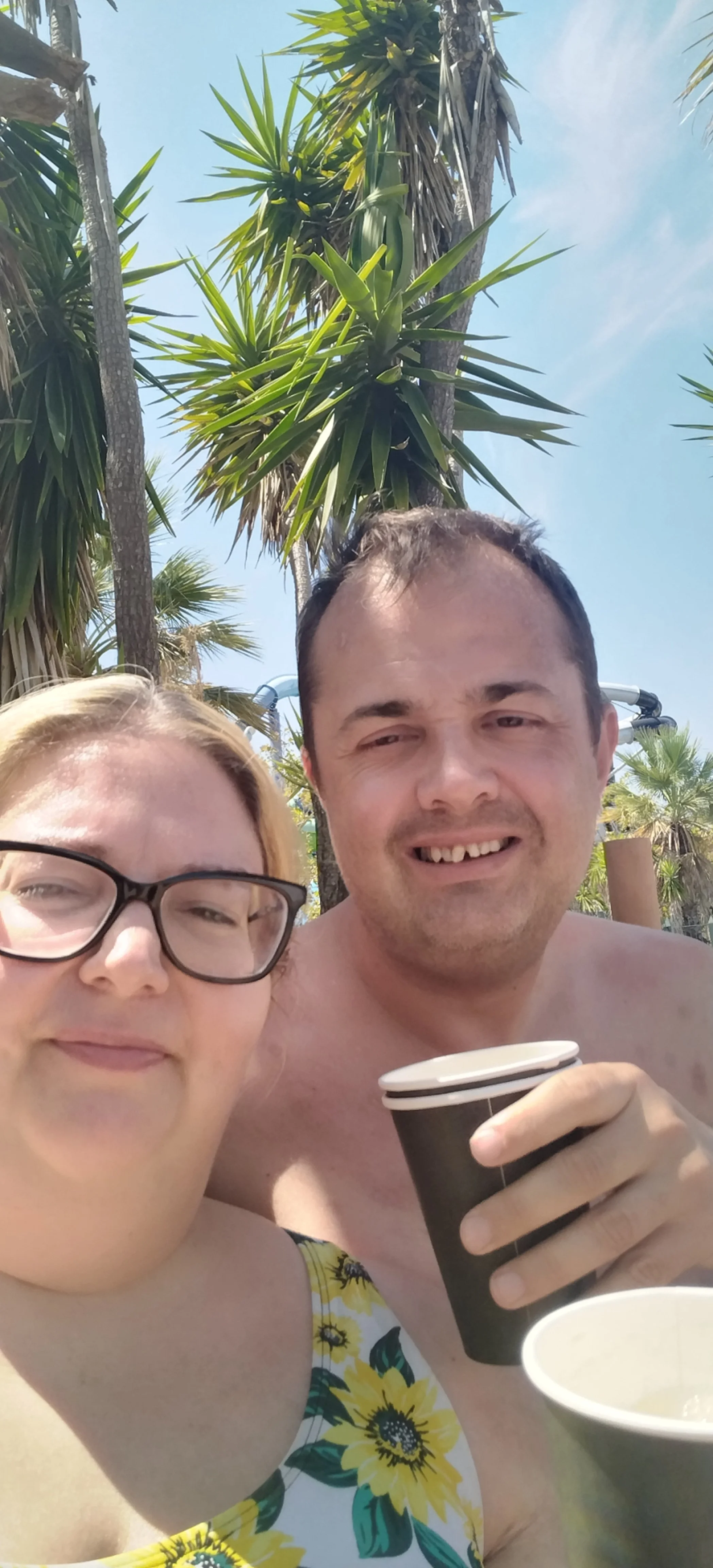 A woman with glasses and blonde hair smiling beside a man with short dark hair, both outdoors in a sunny setting with palm trees, holding cups.