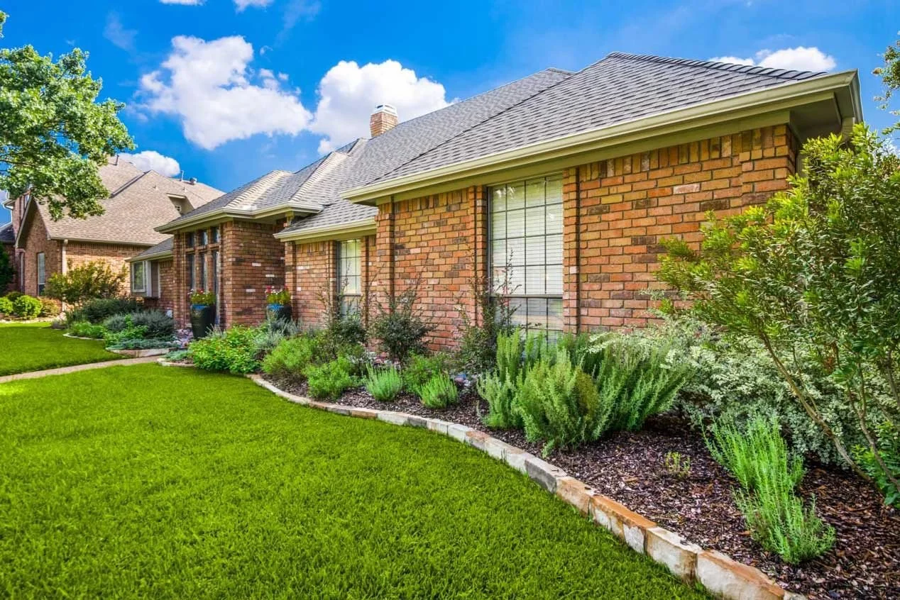 A well-maintained front yard with lush green grass, a flower bed with various plants and bushes, and a brick house under a bright blue sky with a few white clouds.