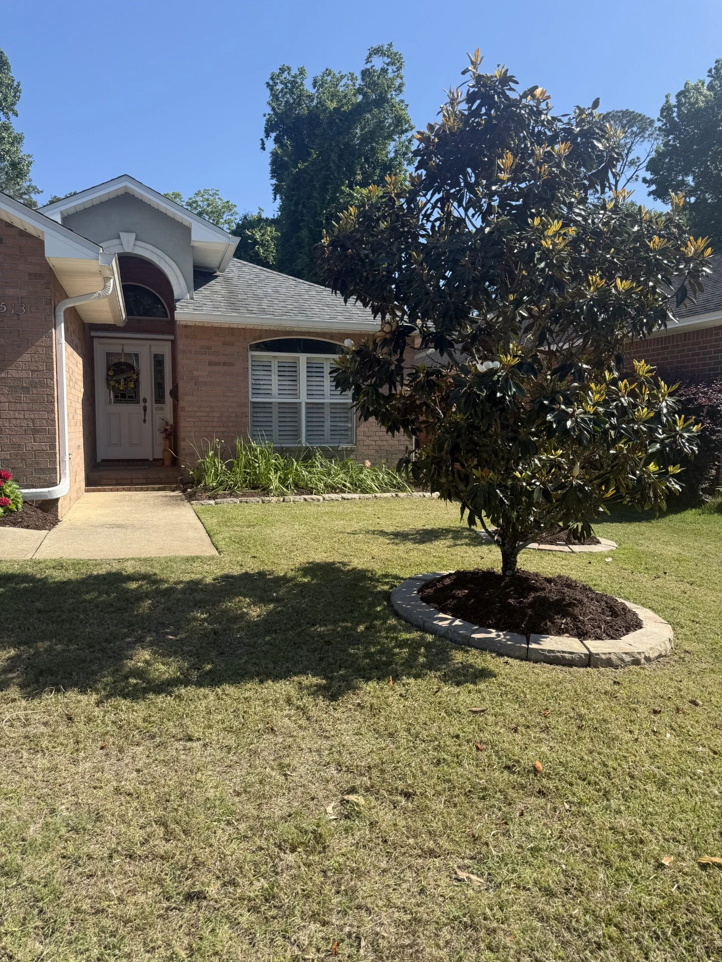 Front view of a brick house with white trim, a small front porch, a large shrub in a circular mulched bed, a grassy lawn, and tall trees against a clear blue sky.