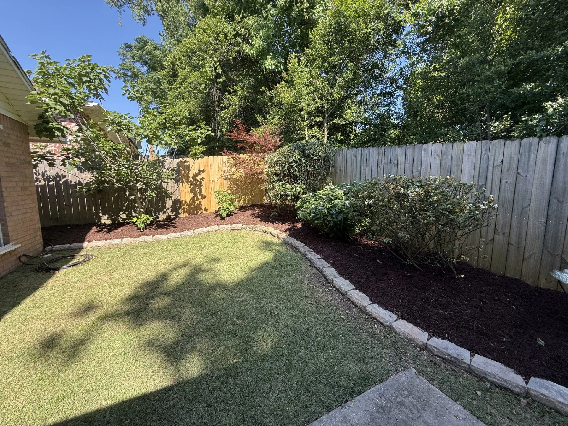 A backyard garden with a green lawn, flower beds bordered with stone edging, and a variety of bushes and trees, surrounded by a wooden fence and a brick house wall on the left.