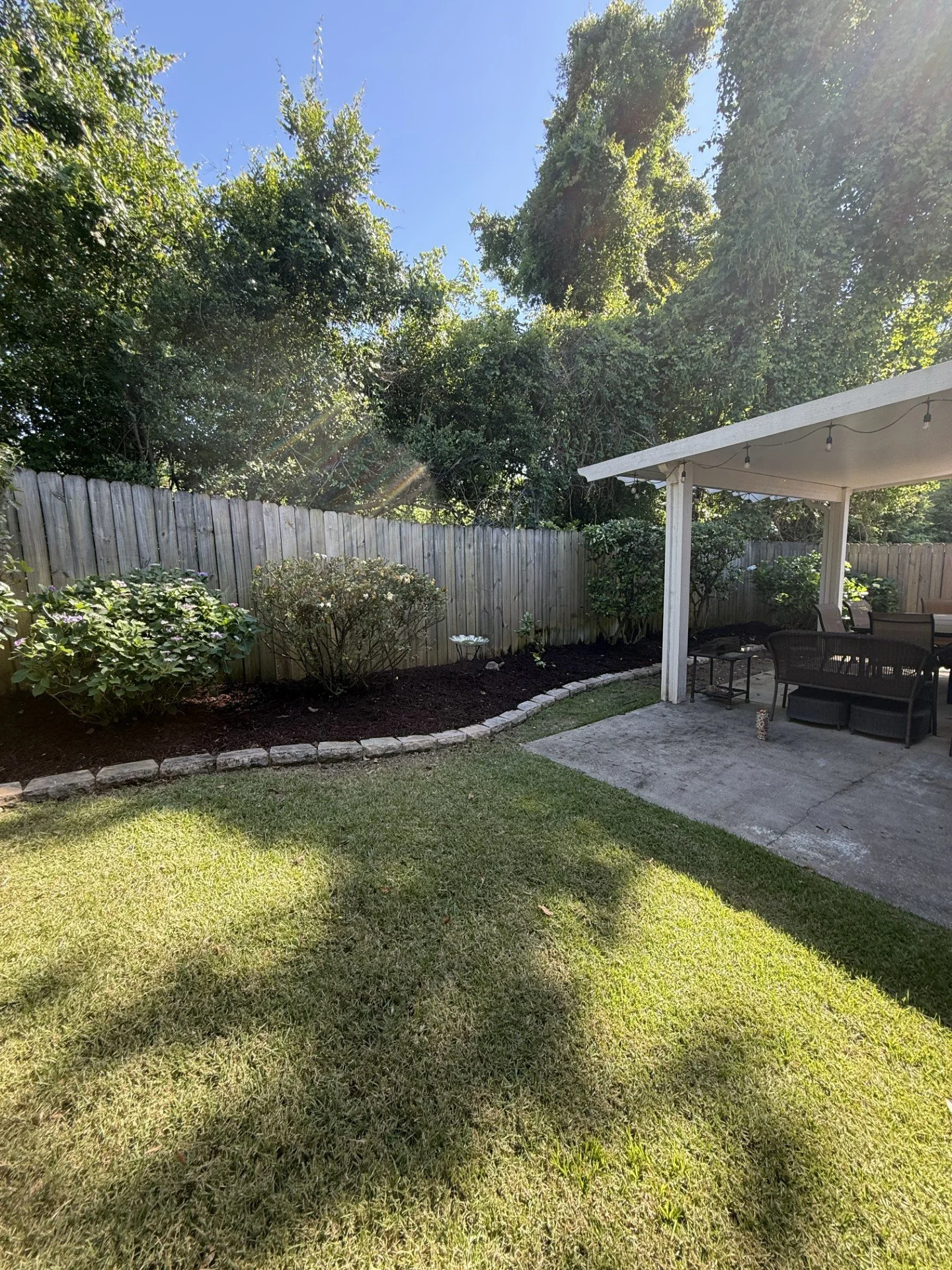 A backyard with a green lawn, a wooden fence, some bushes, a patio with outdoor furniture, and a white covered porch. Trees are visible in the background with a clear blue sky.
