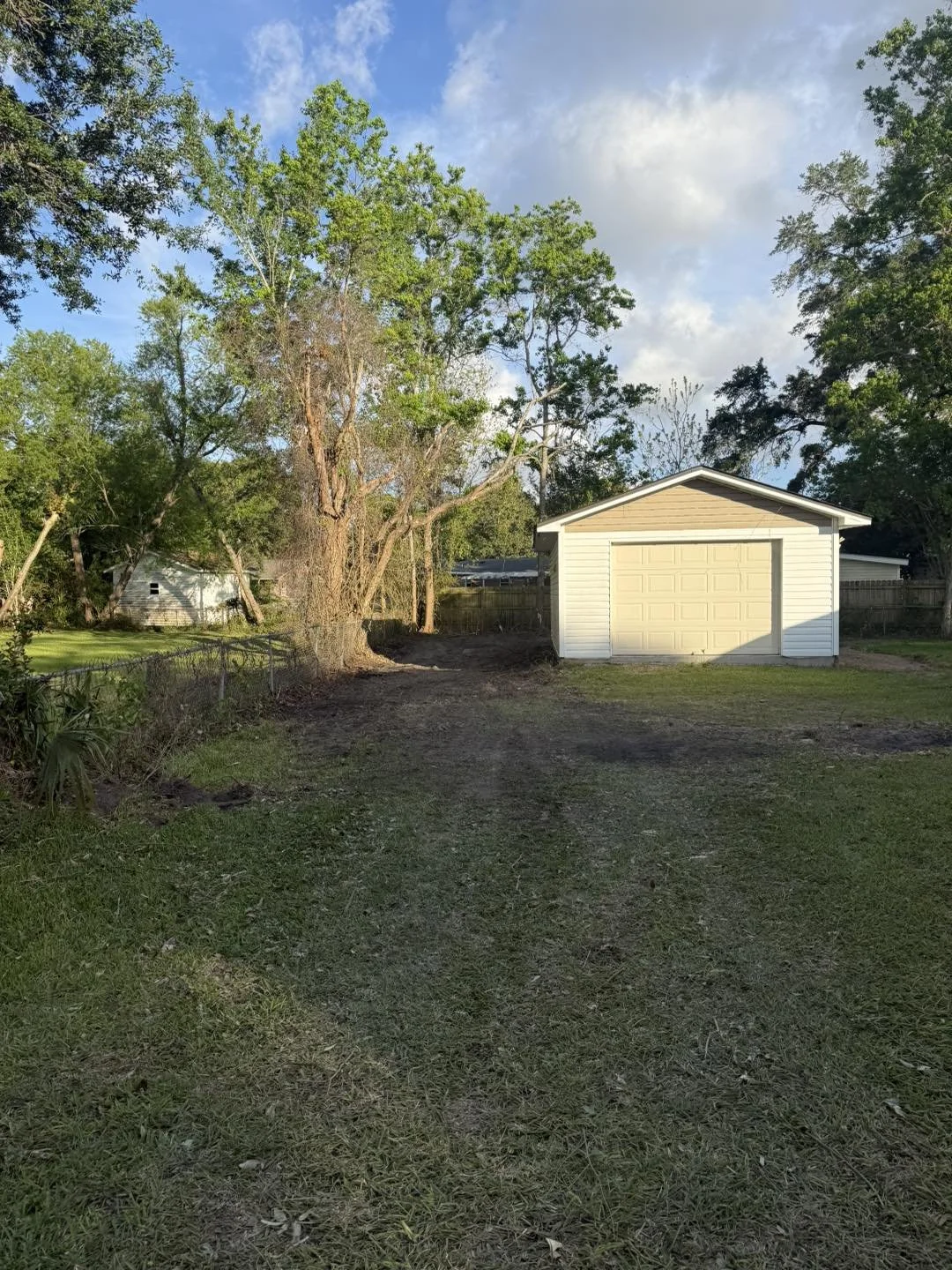A backyard with a large tree, a white garage, and green grass under a partly cloudy sky.