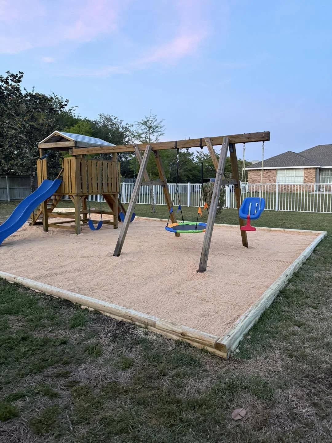 A playground with a sandpit, a wooden play structure with a blue slide, and a swing set with two swings and a baby seat, enclosed by a white fence on a residential yard.