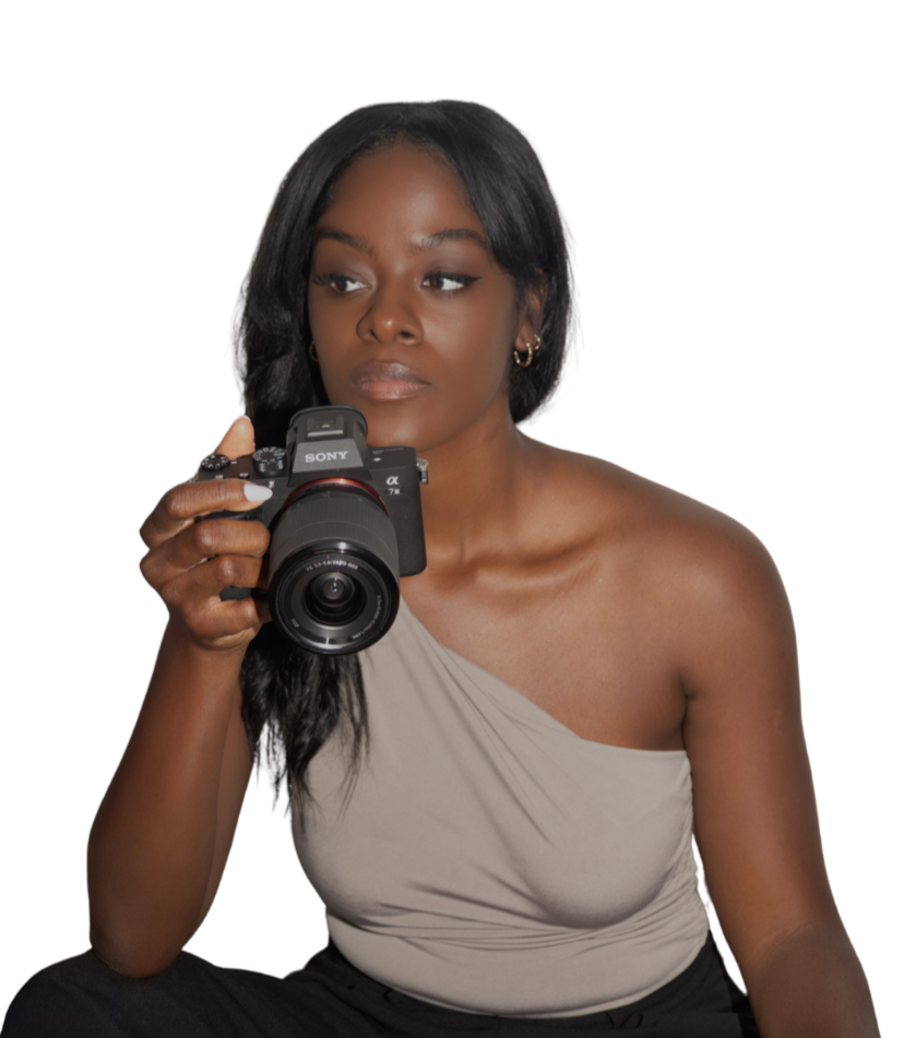 A woman with dark skin and black hair holding a Sony camera, wearing a beige one-shoulder top.