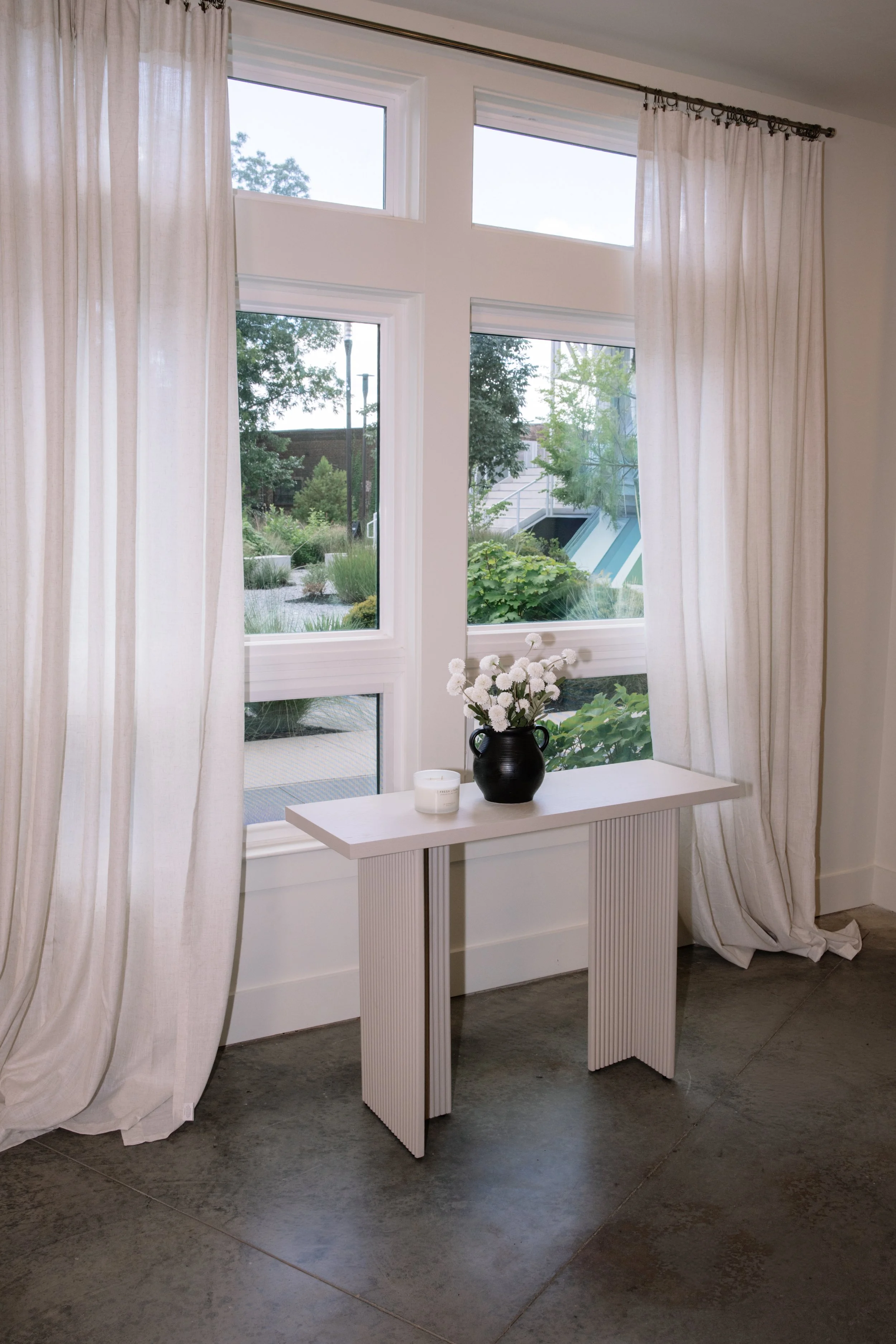Interior view of a room with large windows, white curtains, a small white table with a black vase and white flowers, and a gray floor.