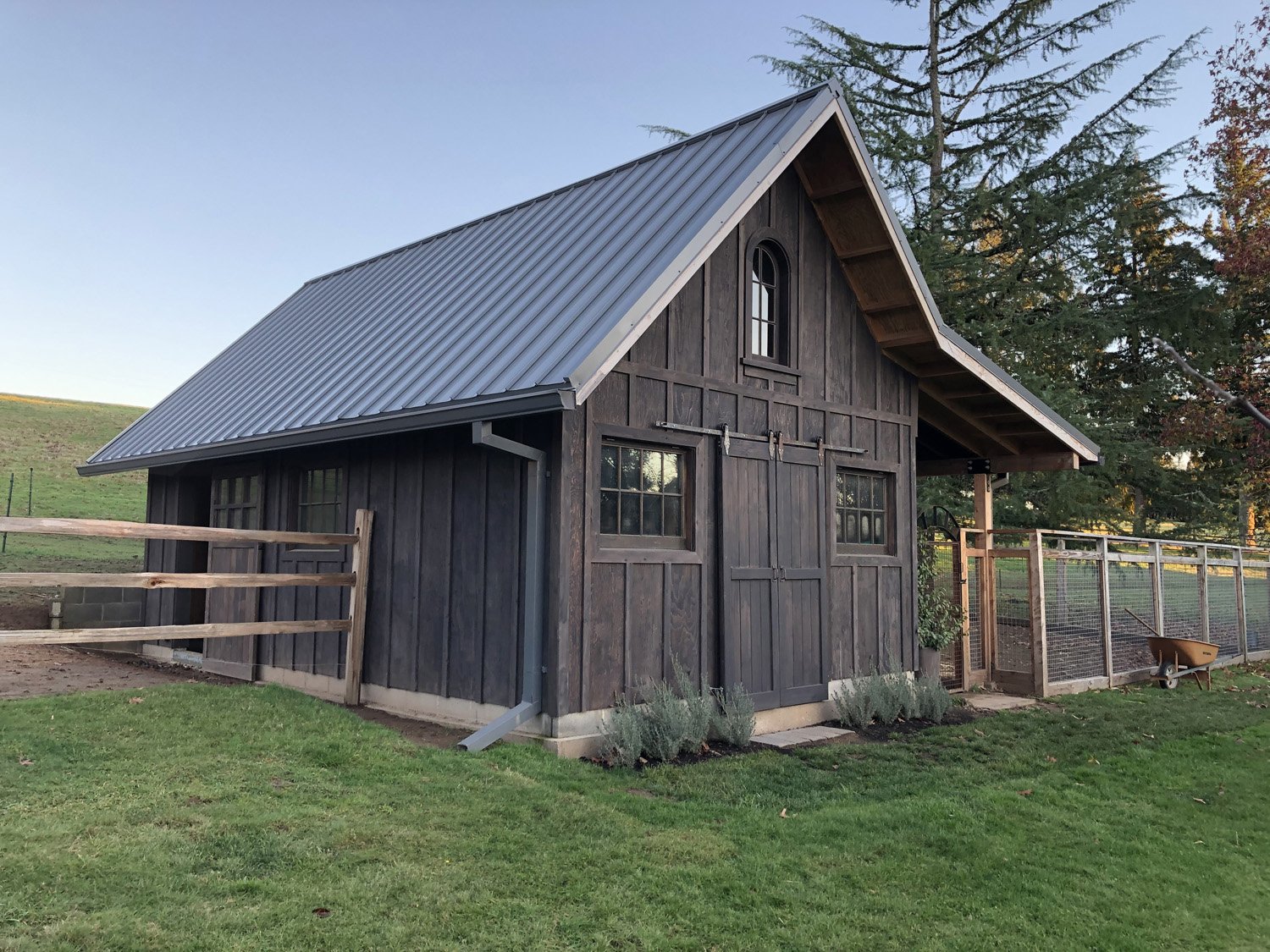 A small black barn with a metal roof and sliding doors, surrounded by a wooden fence and green grass, with trees in the background.