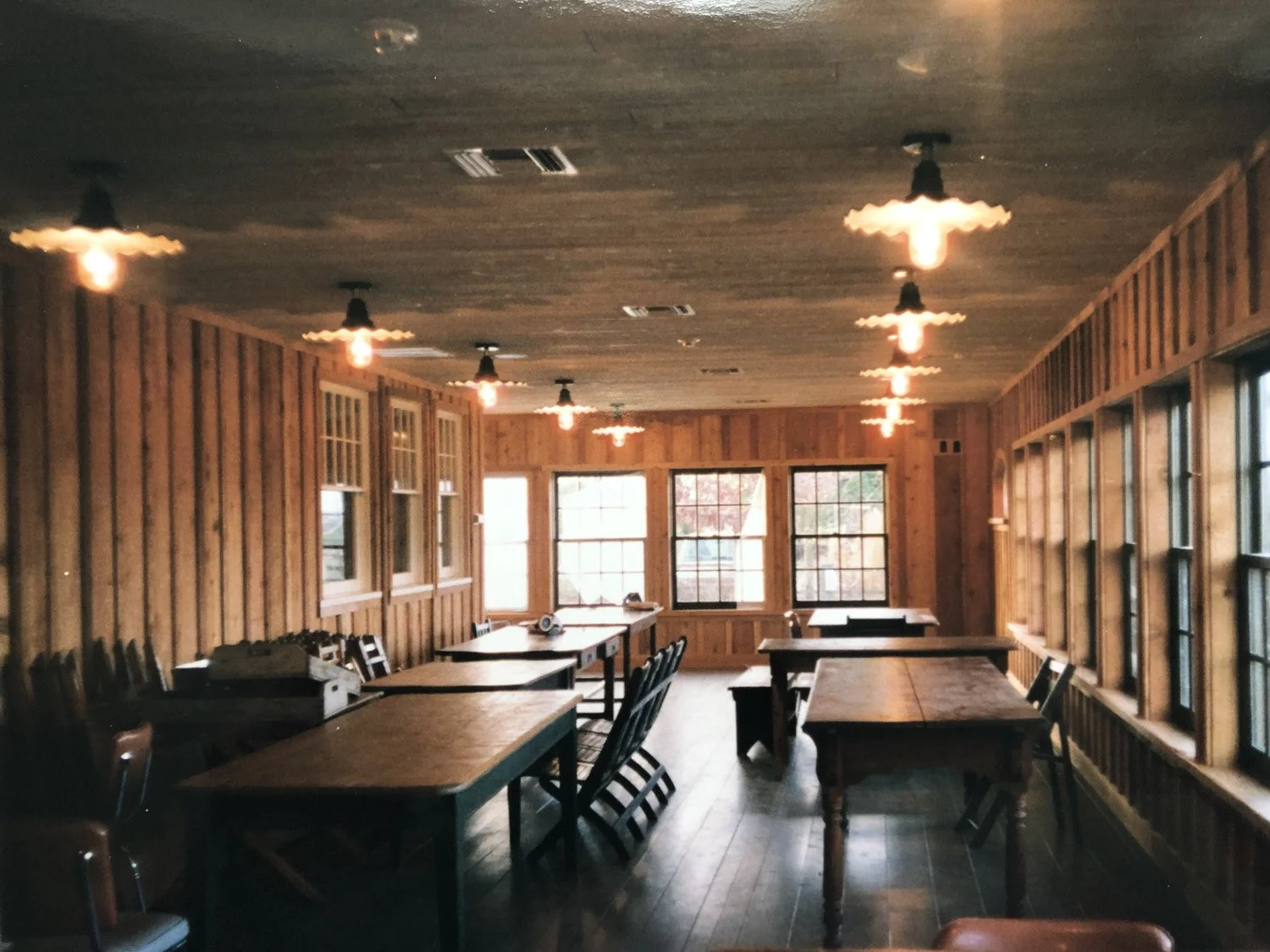 A rustic dining room with wooden walls, multiple windows, and several tables and chairs arranged for seating. The ceiling has pendant lights hanging, and daylight is coming through the windows.