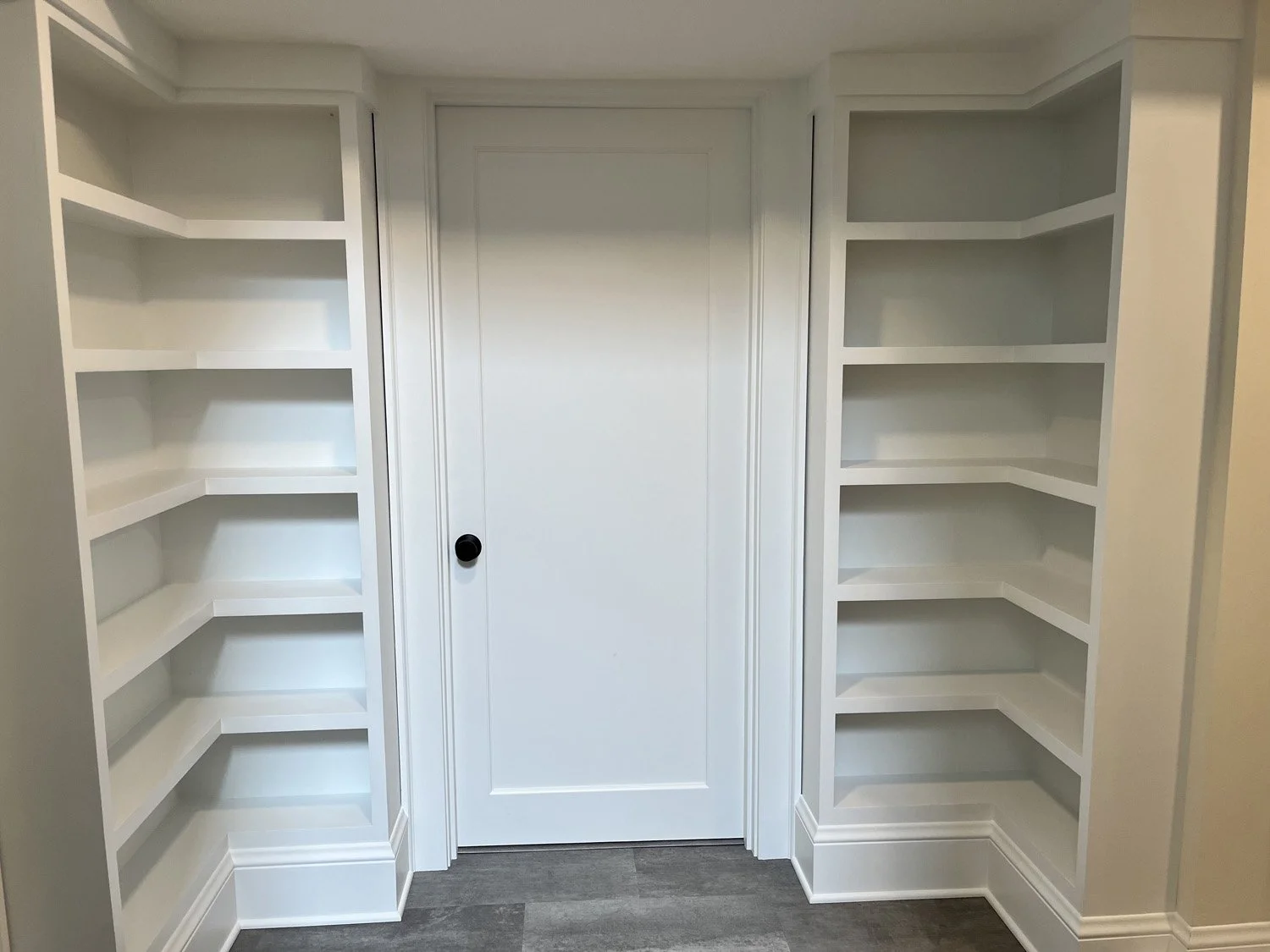 Empty built-in corner shelves with white finish, closed white door with black knob, and gray flooring.