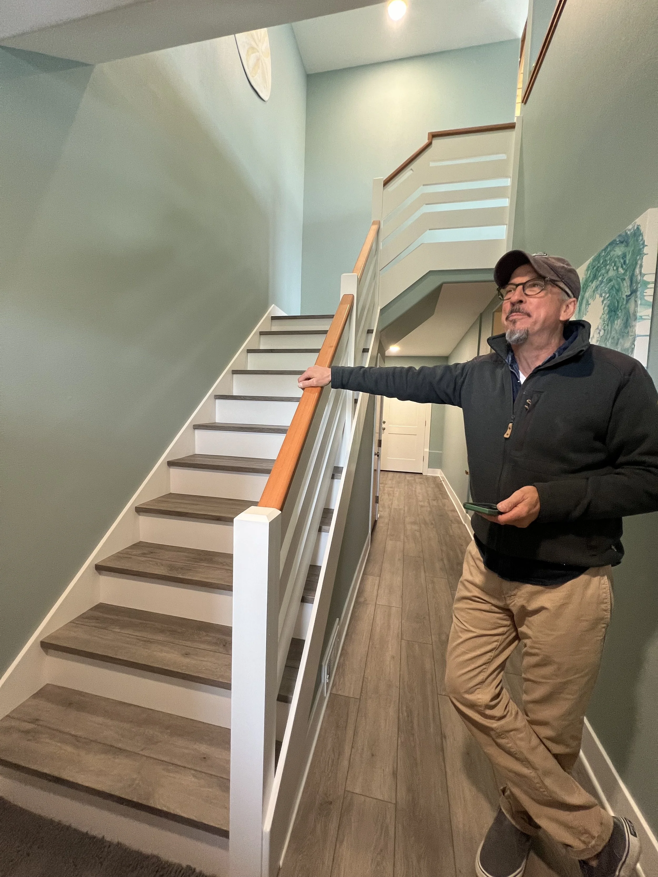 Man standing at the bottom of a staircase inside a house, holding a remote control, wearing glasses, a cap, a black jacket, tan pants, and sneakers.