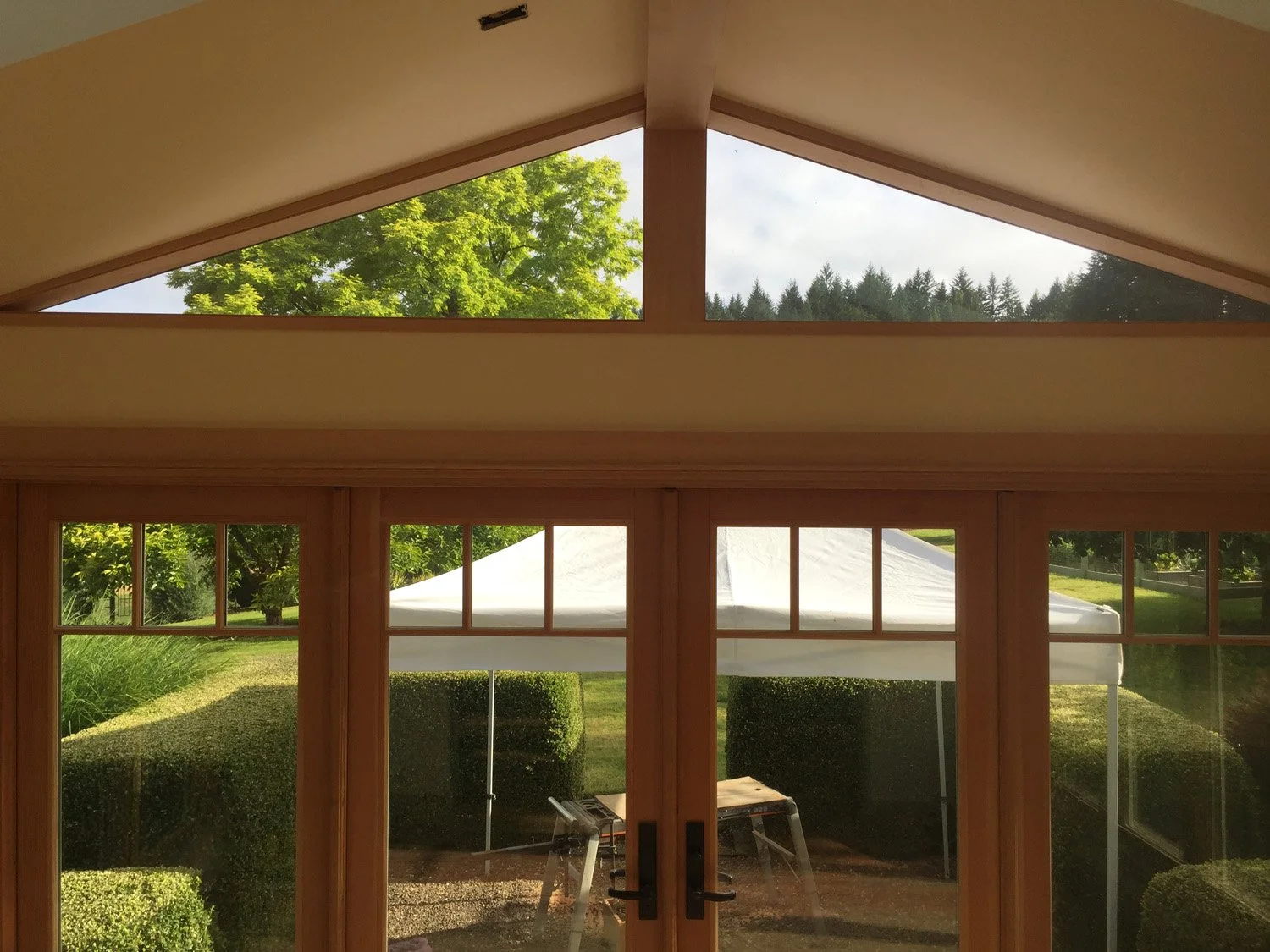 Interior view of a room with large windows and glass doors revealing a backyard with a white tent, lush green lawn, hedges, trees, and a mountain range in the background.