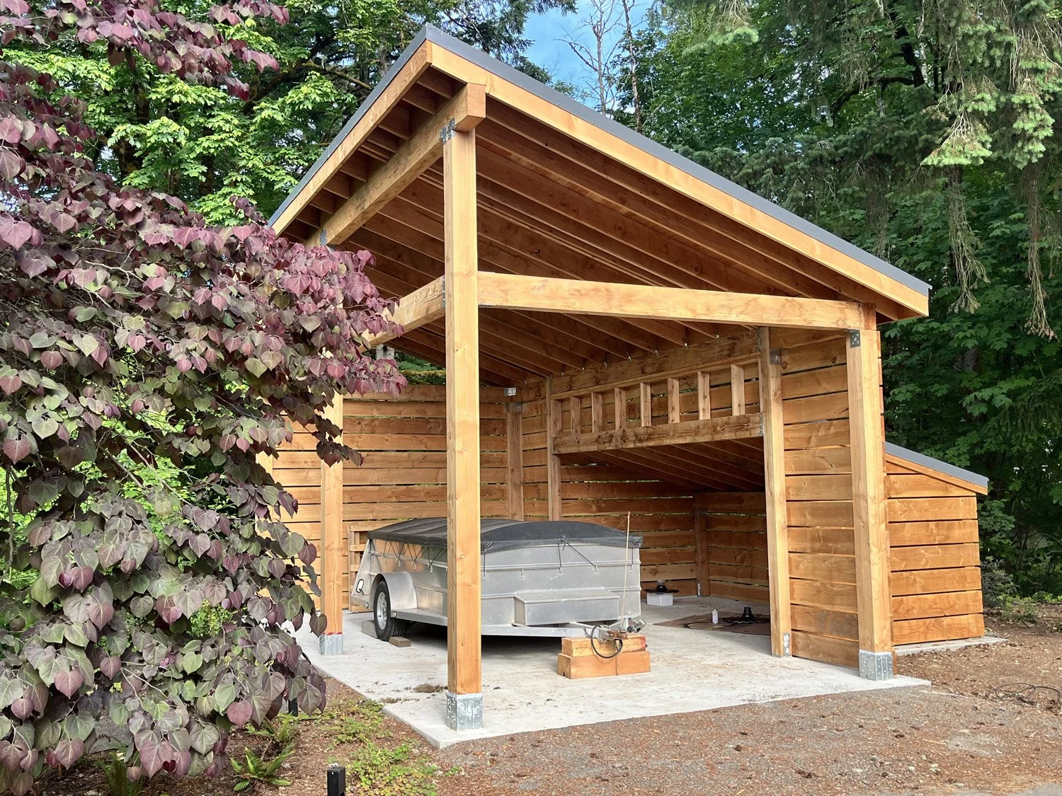 A wooden carport structure with a metal roof, situated outdoors near trees and plants. Under the carport, there is a boat trailer with a small boat attached, on a concrete pad.