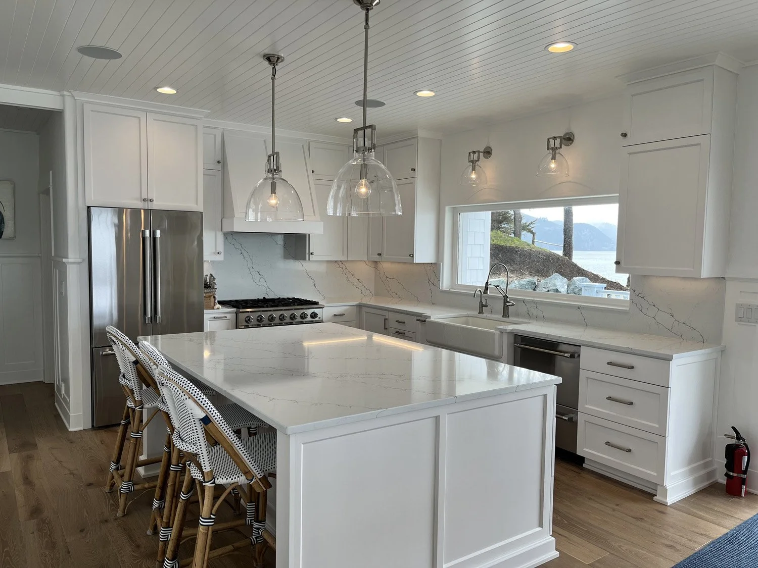 Modern white kitchen with marble countertops, stainless steel refrigerator, oven, and large window overlooking a scenic landscape with trees and water.
