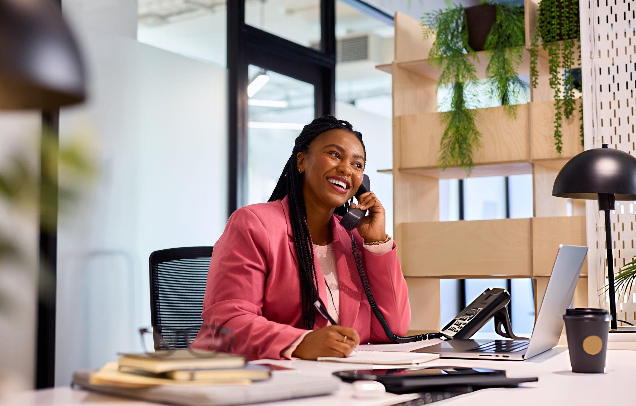 A woman with braided hair and a pink blazer smiling while talking on a landline phone at a modern office desk with a laptop, coffee cup, and notebooks, with green plants and decorative wooden shelves in the background.