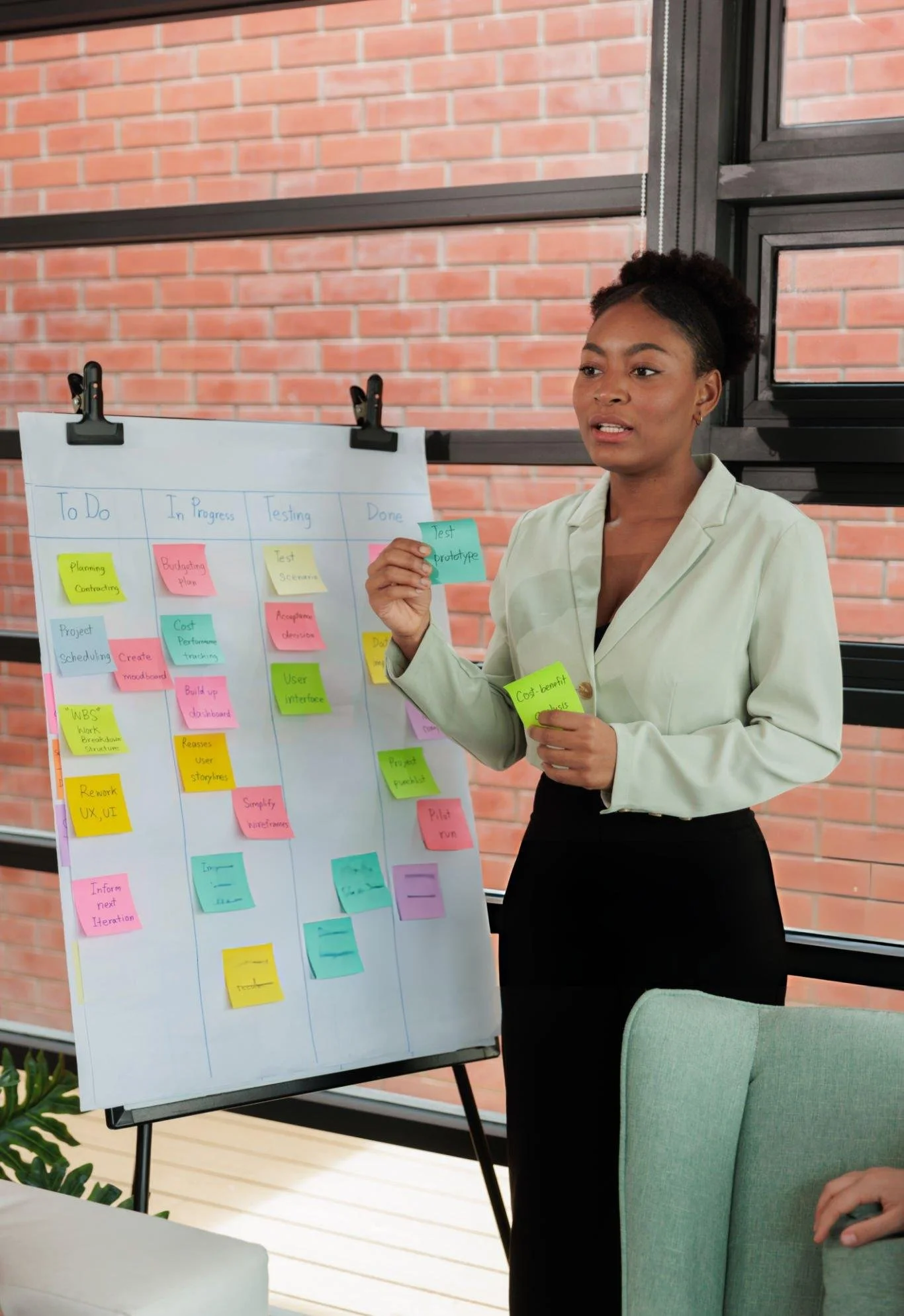 A woman in a beige blazer holding a sticky note, standing in front of a large chart with colorful sticky notes organized into columns labeled 'To Do,' 'In Progress,' 'Testing,' and 'Done' in an office setting with brick walls and large windows.