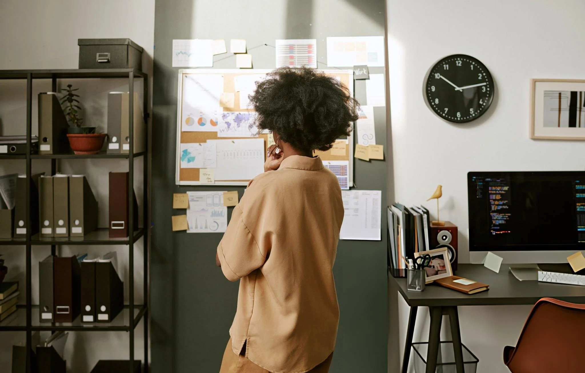 A person with curly hair, dressed in a beige shirt, standing in an office, looking at a bulletin board filled with charts, notes, and maps, with a clock on the wall showing 2:49.