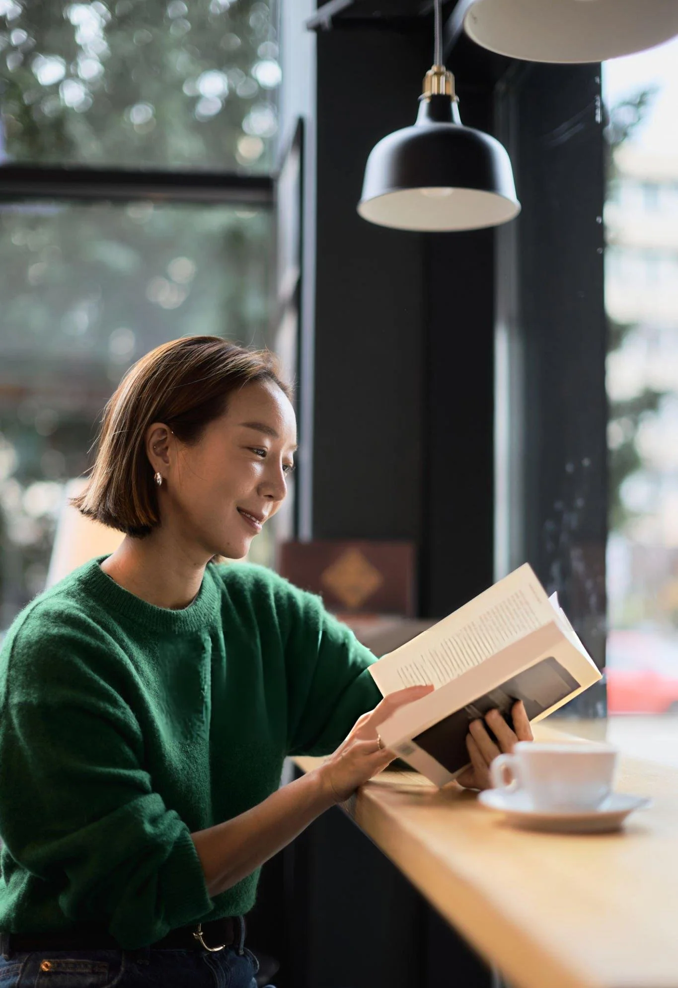 A young woman with short brown hair wearing a green sweater and earrings sits at a cafe counter, reading a book. There are cups on the counter, and who appears to be a coffee shop or cafe setting with large windows and natural light.