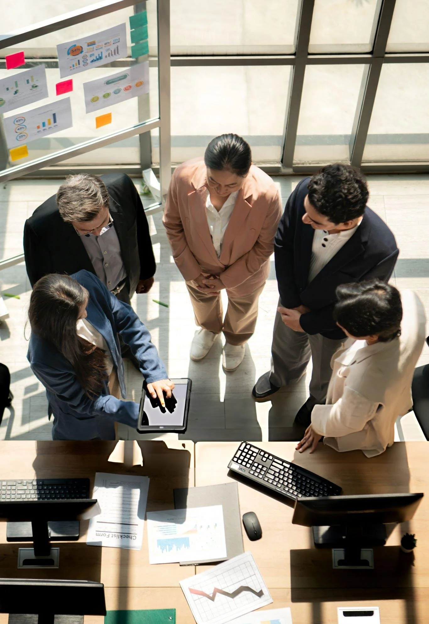 Business meeting with six professionals gathered around a table with computer keyboards, documents, and charts, in a bright office with large windows.