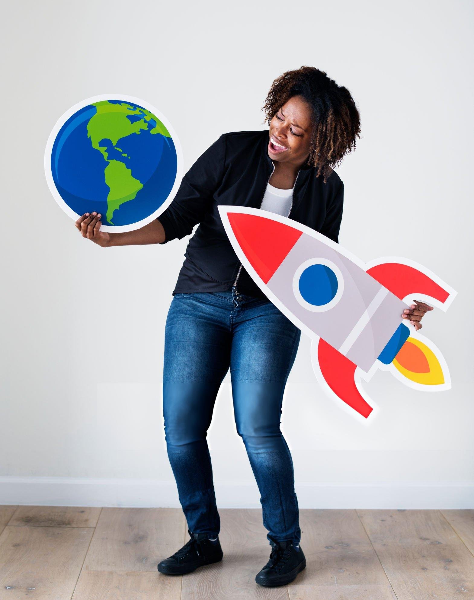 A woman holding a paper cutout of the Earth in front of her and a rocket-shaped paper cutout in her other hand, standing on a wooden floor against a plain white background.