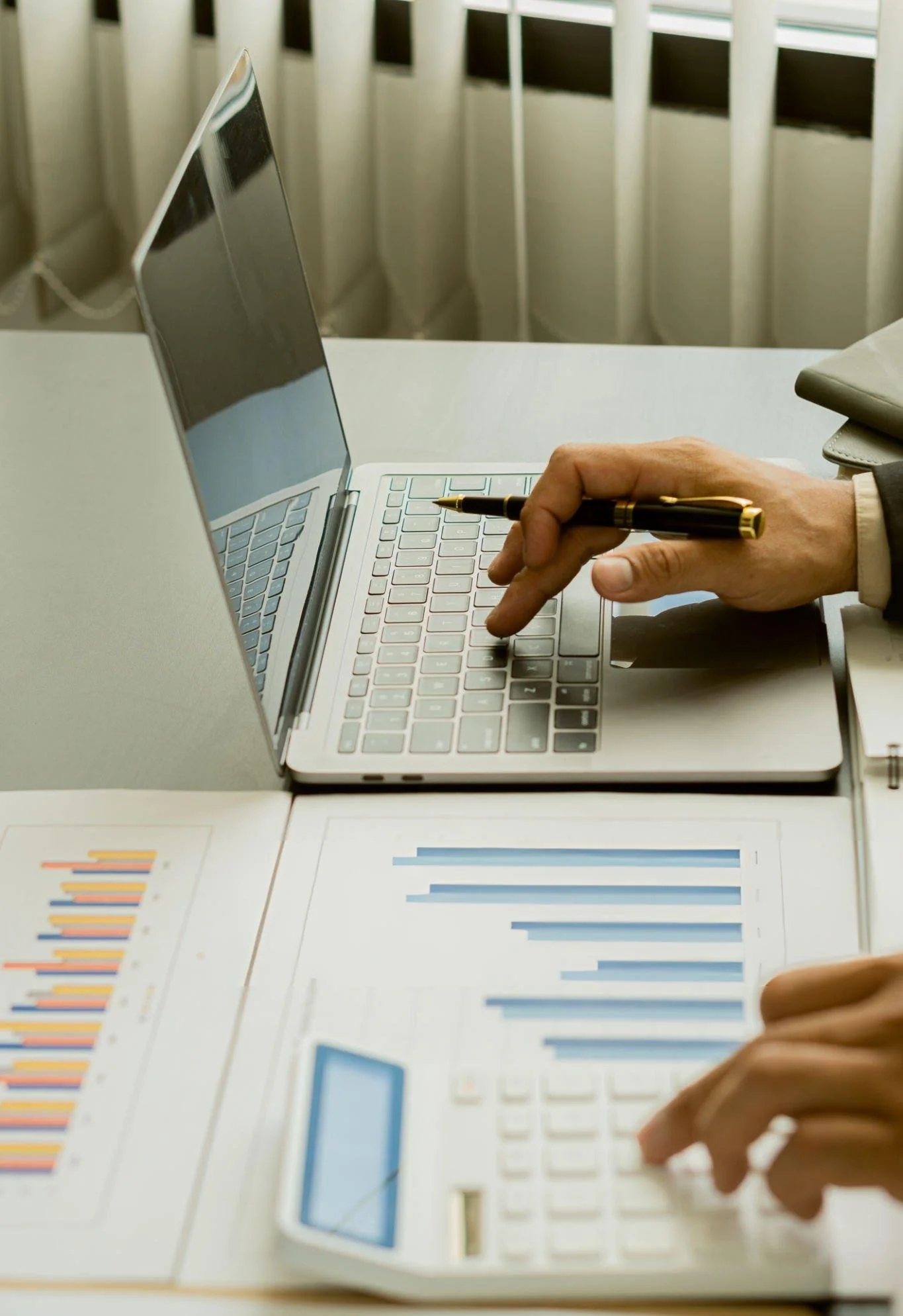 A person's hand holding a black and gold pen over a laptop keyboard, with documents featuring bar graphs and charts on the table.