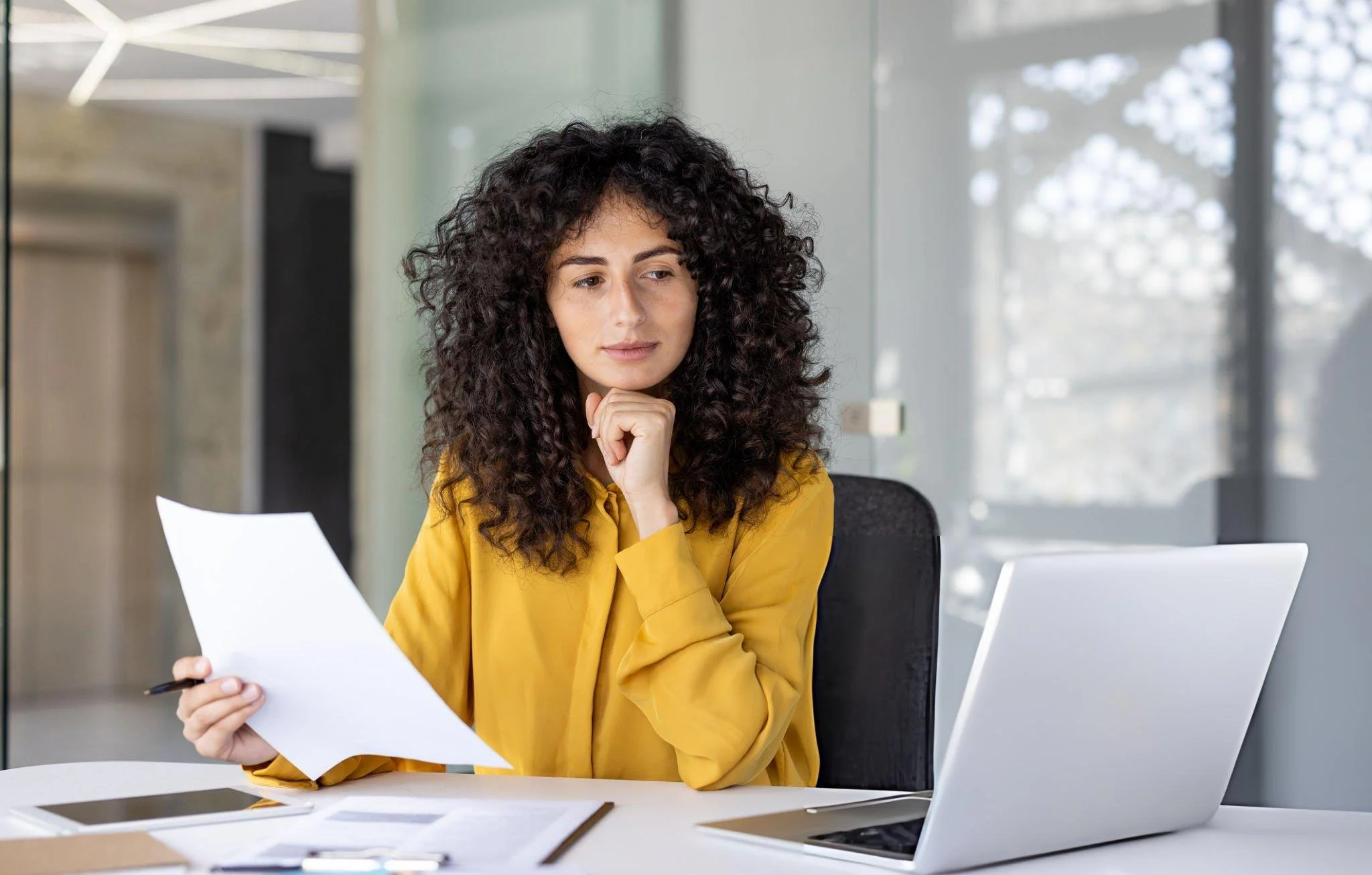 A woman with curly hair and a yellow blouse sitting at a desk, looking at papers and holding a pen, in an office with large windows.
