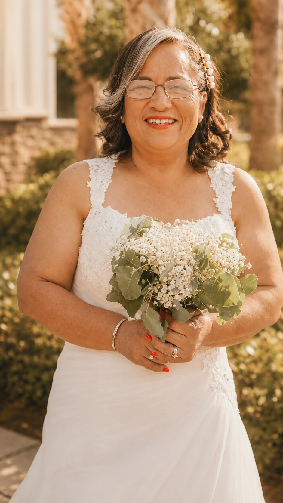 A smiling woman in a white wedding dress holding a bouquet of white flowers outdoors.
