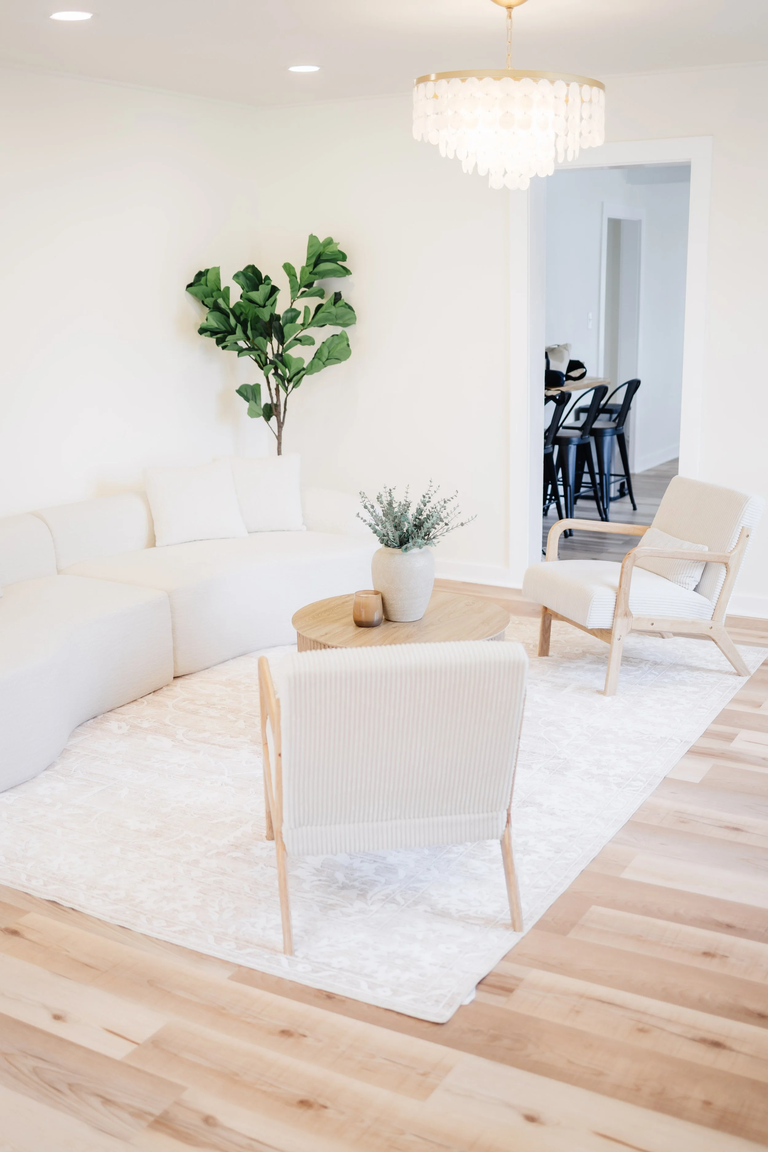 A bright, modern living room with a white curved sofa, a round wooden coffee table, and two white armchairs with wooden frames. There are green plants, a white rug, and a chandelier with shell-like decorations hanging from the ceiling. A doorway reveals a kitchen area with black barstools and a counter.