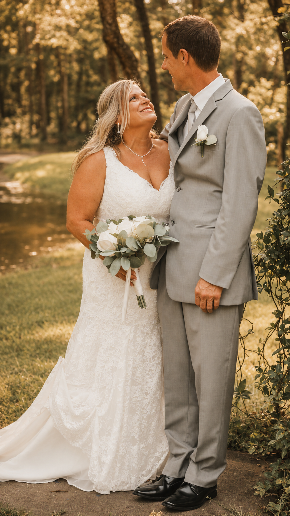 A bride and groom standing outdoors on their wedding day, surrounded by trees, with the bride holding a bouquet of white flowers and wearing a white lace wedding dress, while the groom is dressed in a light gray suit, both smiling at each other.