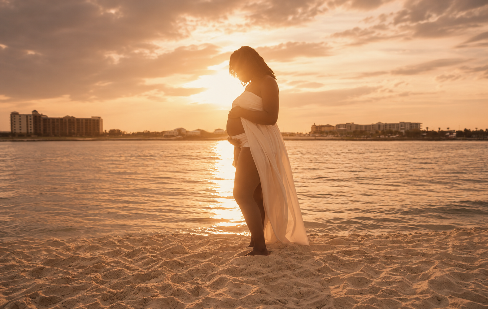 A silhouette of a pregnant woman standing on a sandy beach at sunset, wearing a flowing white dress, with buildings in the background and water reflecting the sunset