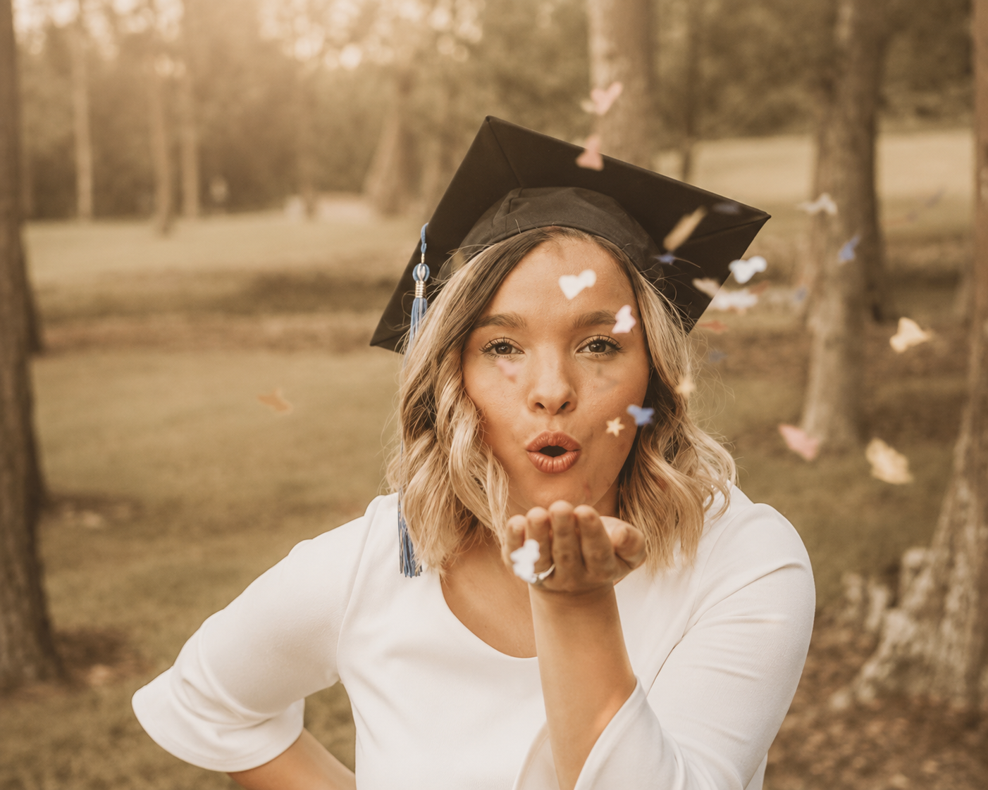 A woman in a white jacket blowing confetti with forest trees in the background, wearing a graduation cap.