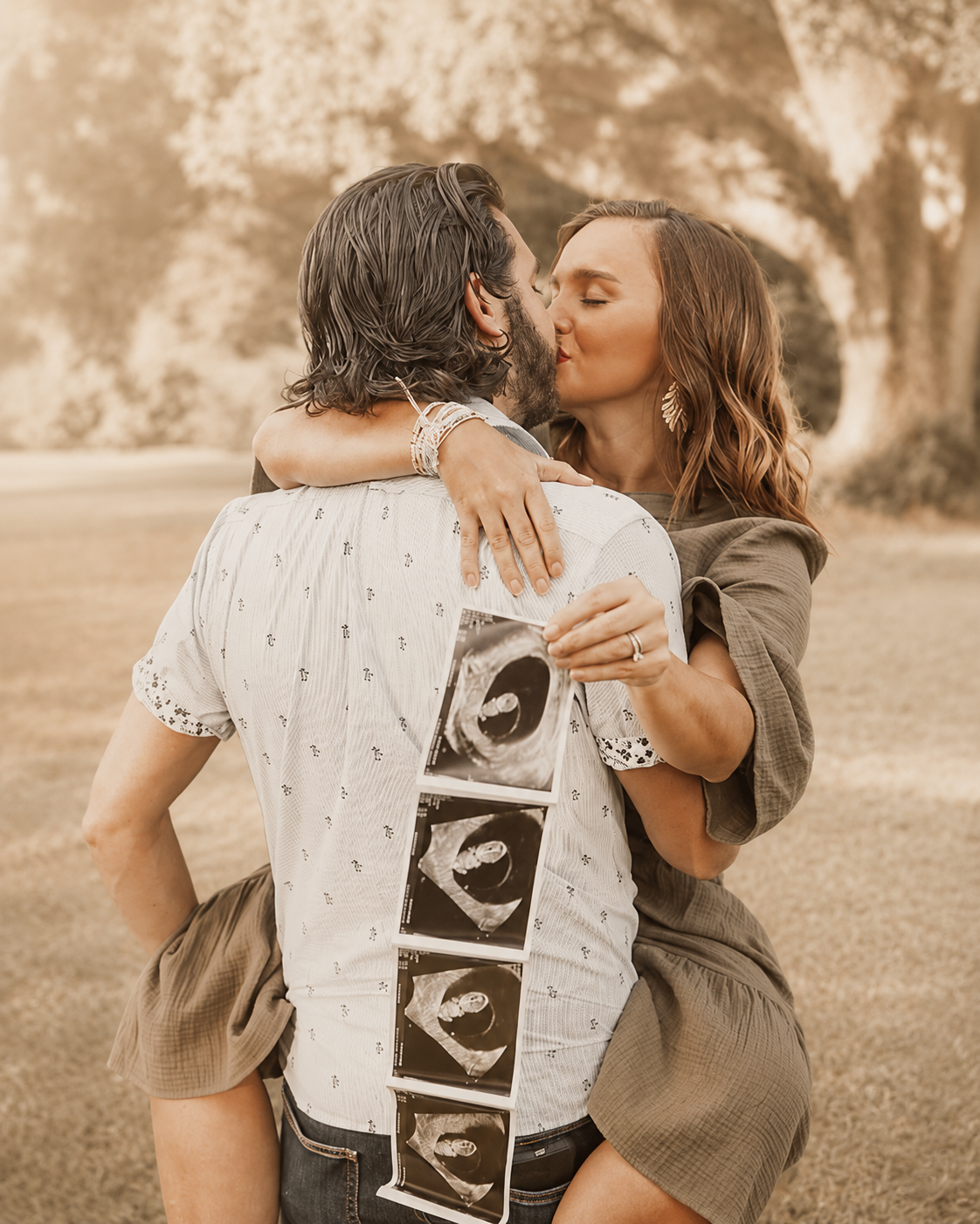 A couple kissing outdoors, holding ultrasound pictures showing an unborn baby.