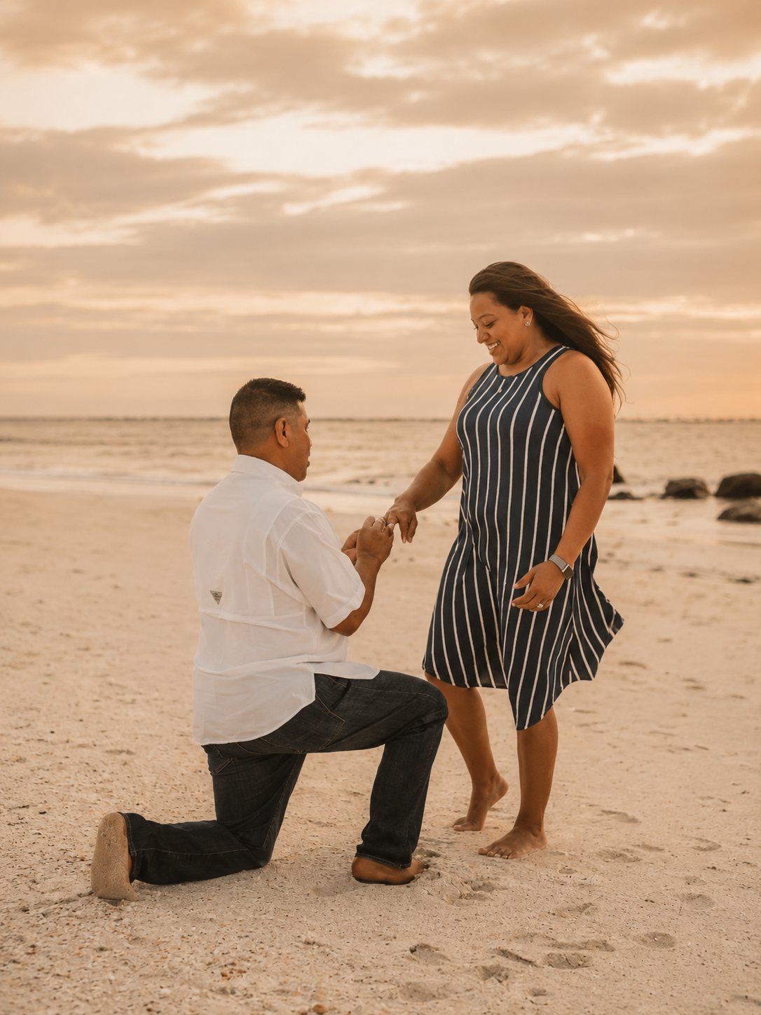 A man kneeling on the beach during sunset, holding hands with a woman standing, both smiling.