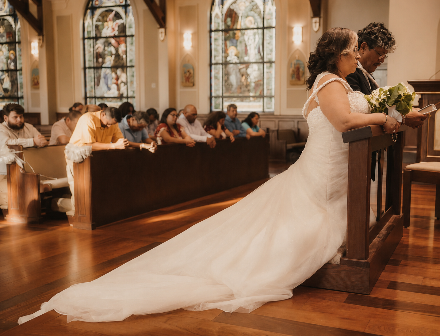 A bride praying with her parents in a church during a wedding ceremony, with guests seated in pews and stained glass windows in the background.