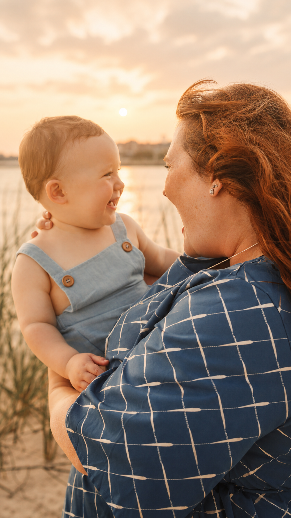 A woman with red hair smiling and holding a young child at sunset near a body of water.