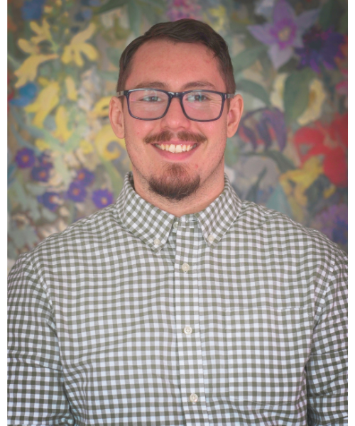 A young man with short dark hair, glasses, and a goatee, smiling, wearing a green and white checkered button-up shirt, standing in front of a colorful floral background.