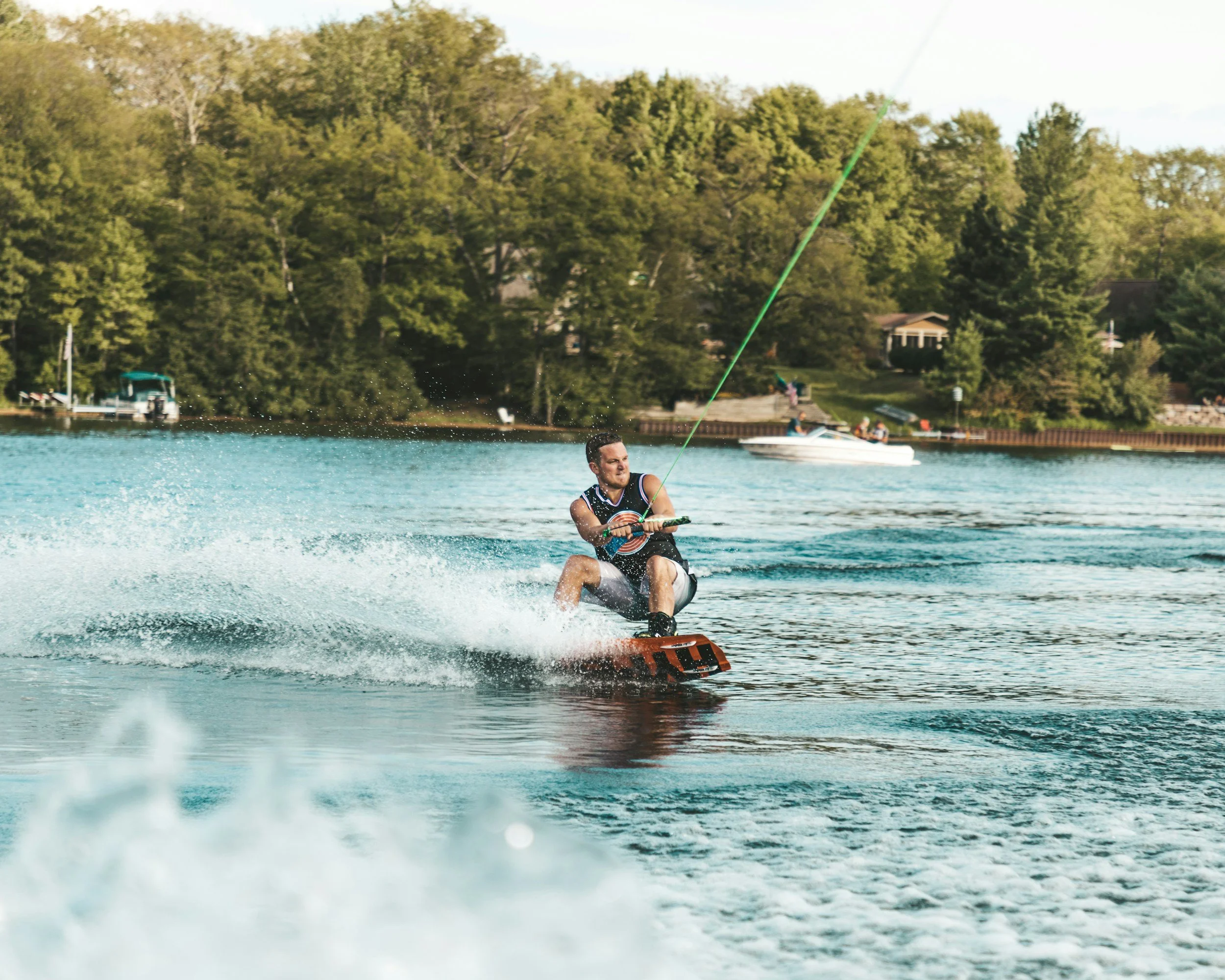 Man wakeboarding on a lake, holding a green rope handle, with trees and houses in the background.